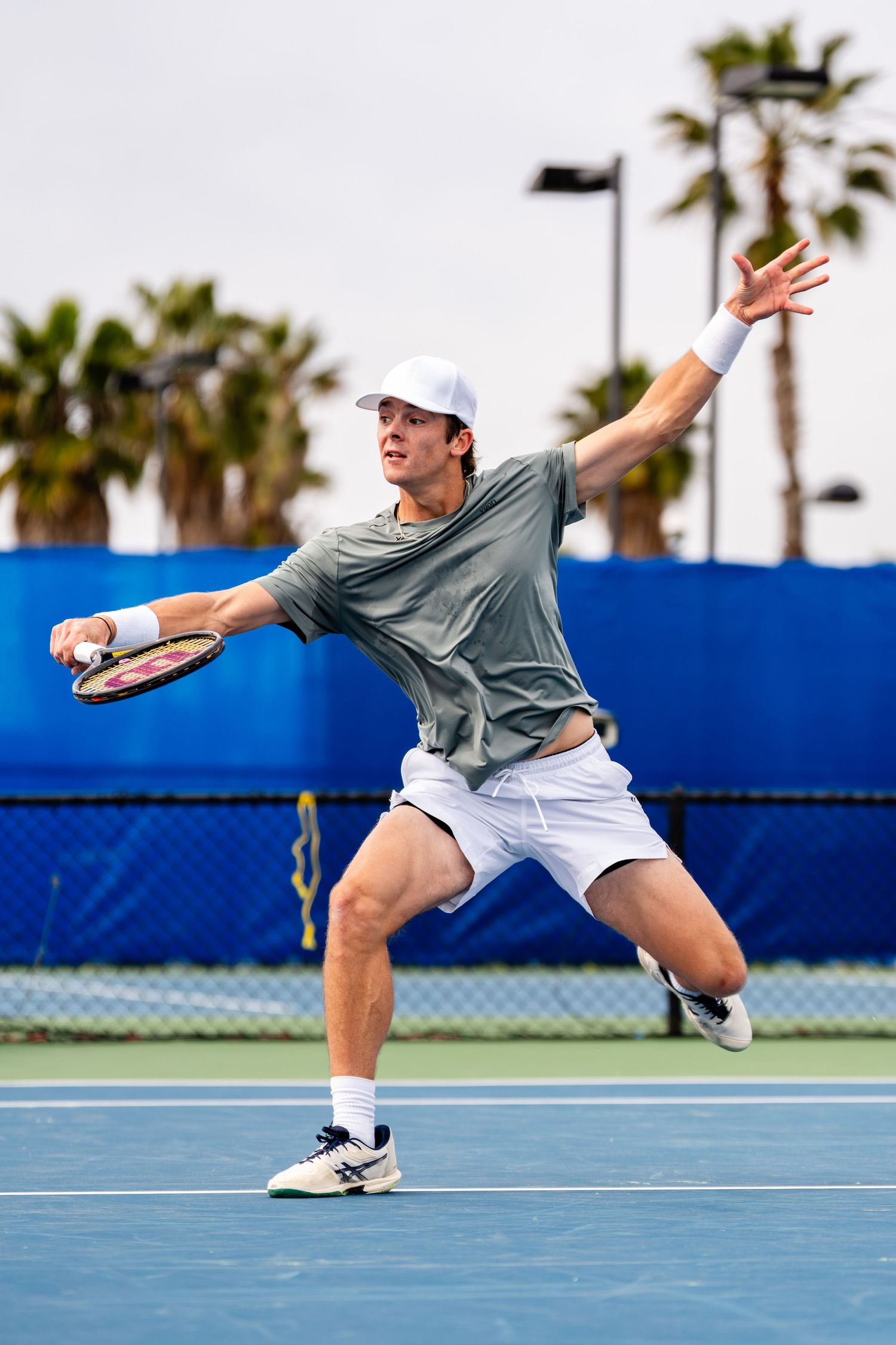 Male tennis player lunges to hit forehand shot on outdoor blue hard court with palm trees background.