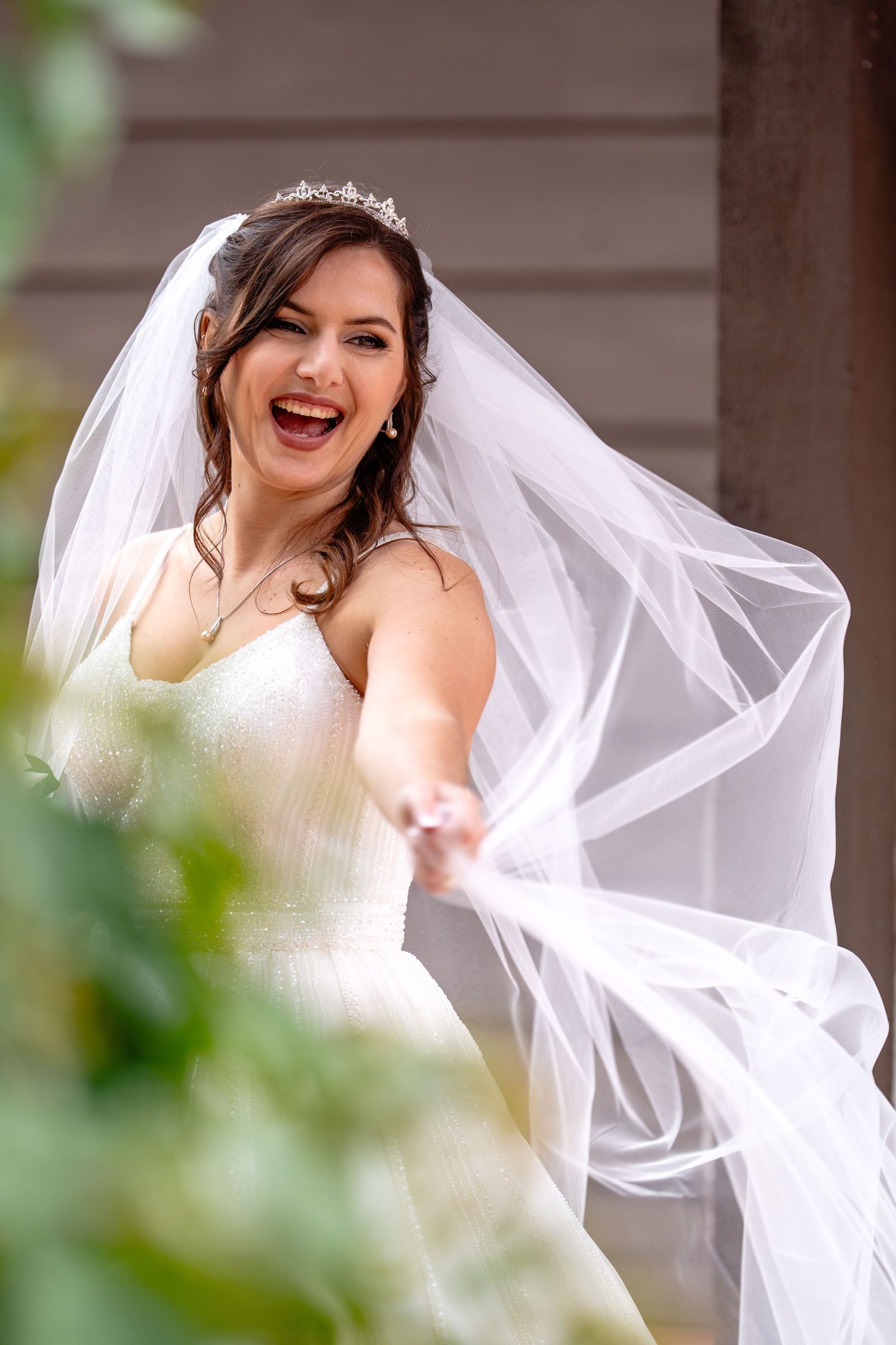 Joyful bride in white strapless gown and tiara laughs as her flowing veil billows in the wind.