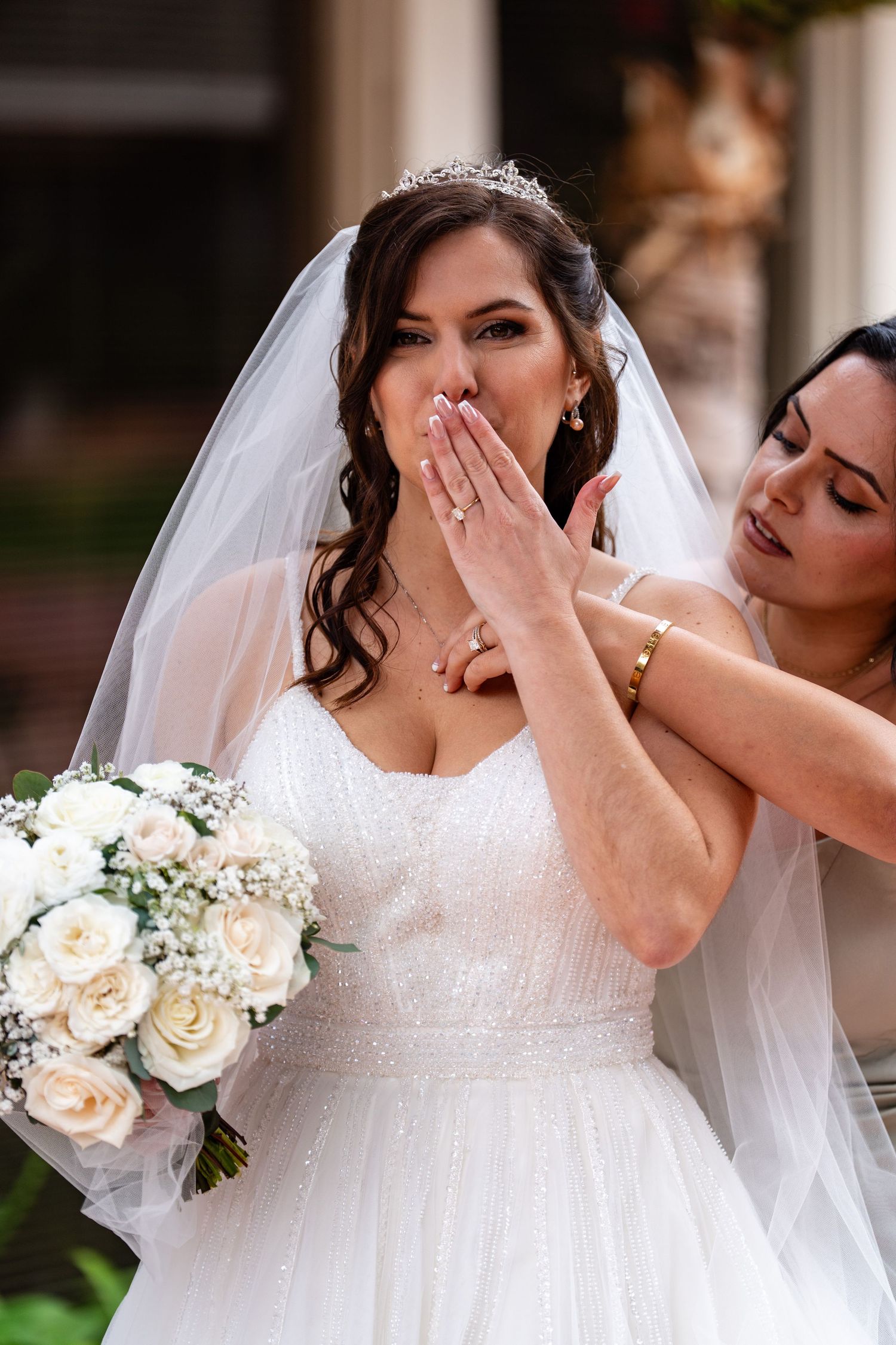 Bride in white gown and veil holds roses bouquet while bridesmaid gently touches her face during wedding preparation.