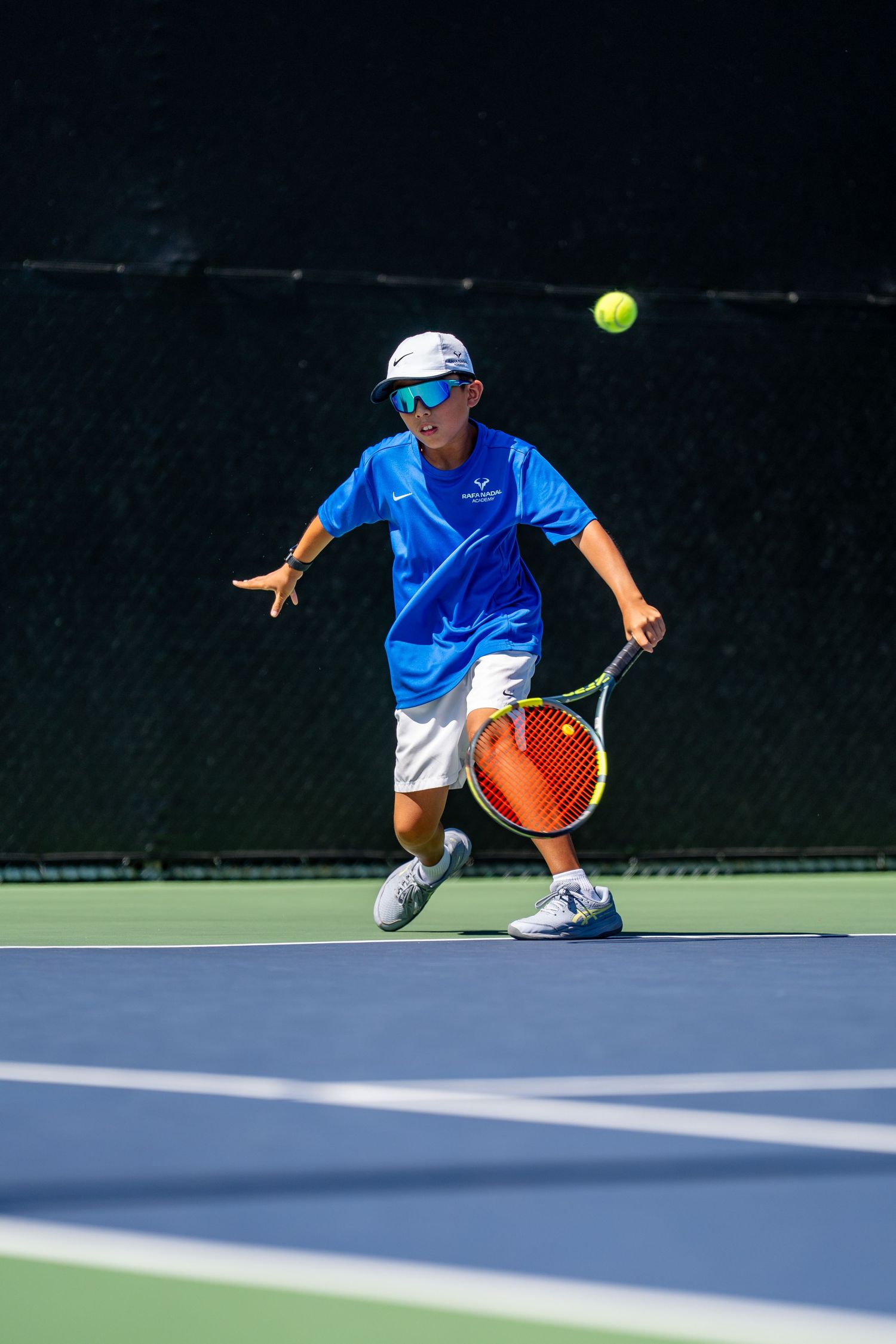 Young boy in blue shirt and cap playing tennis, reaching to hit a yellow ball with an orange racket on a sunny court.