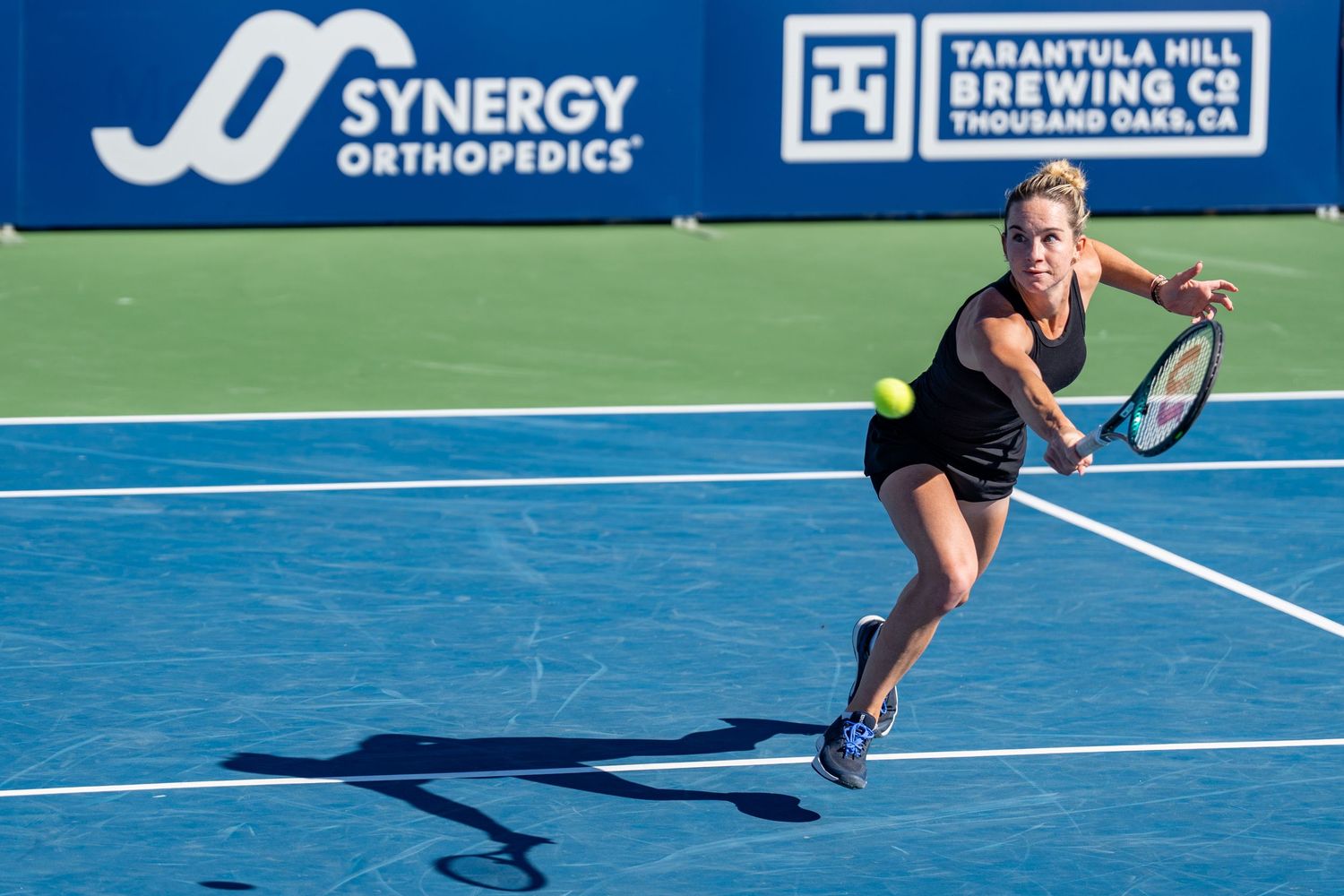 Female tennis player in black outfit sprinting to return a shot at the Synergy Orthopedics sponsored tennis tournament.
