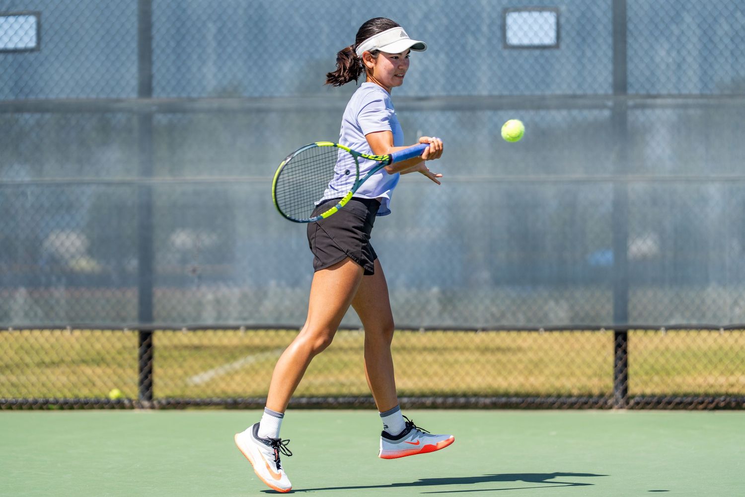 Young female tennis player hits a forehand shot during an outdoor match on a sunny day.