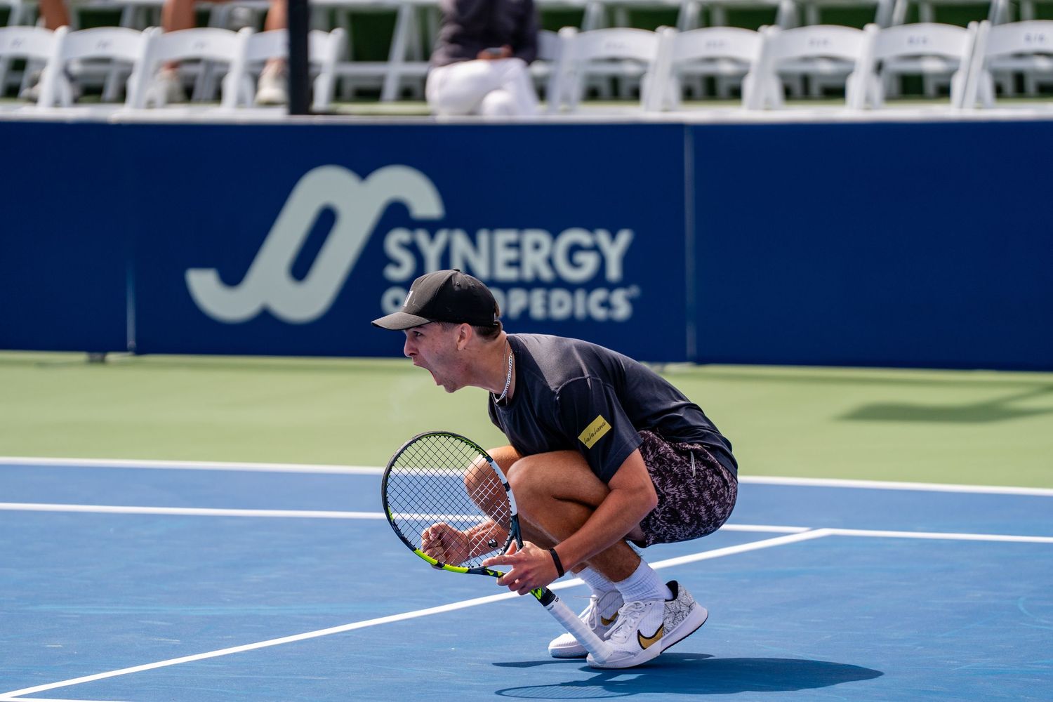 Tennis player crouches low on a blue hard court near a Synergy Orthopedics banner during a match.
