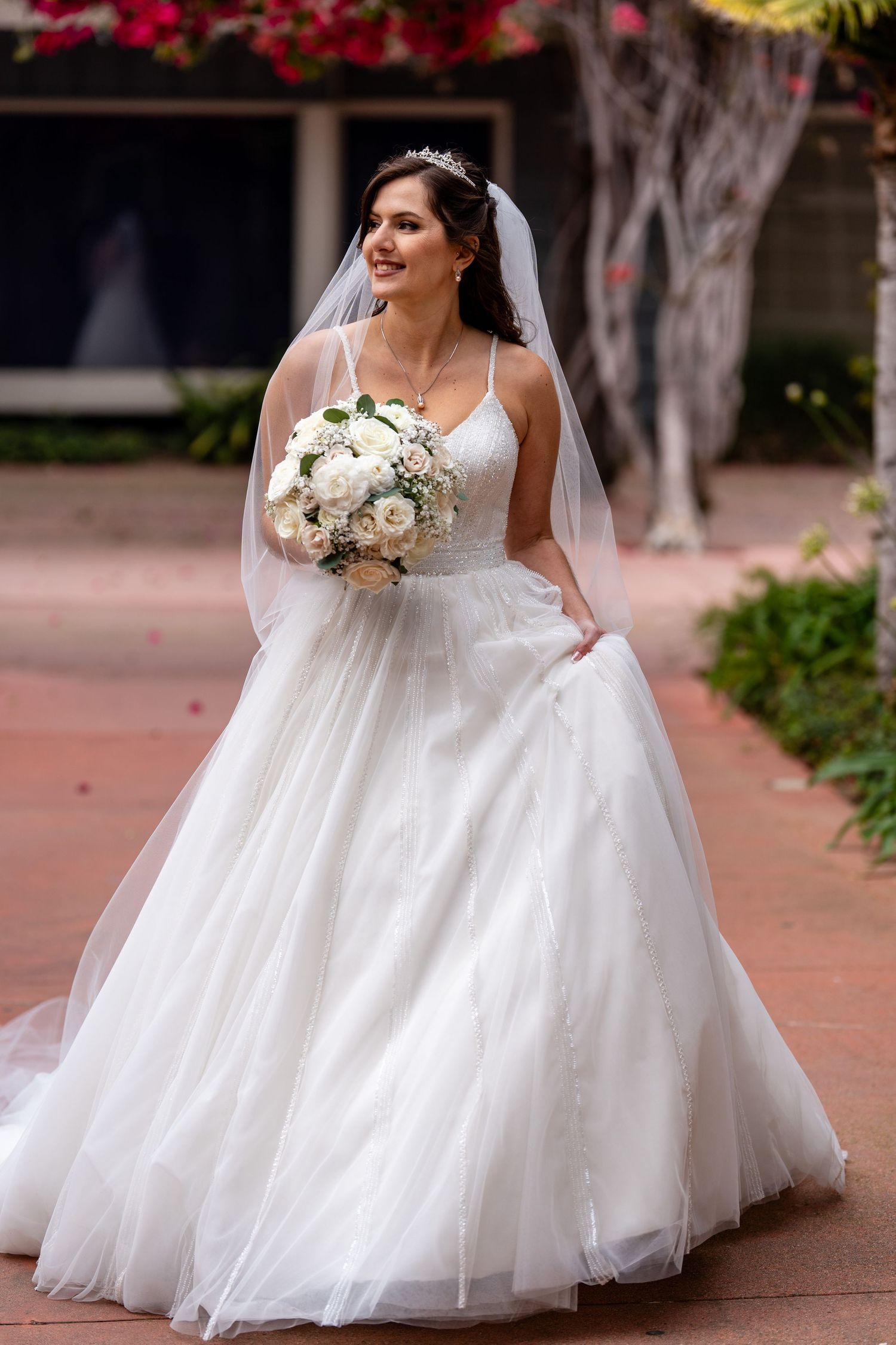 Smiling bride in a white ball gown with veil and tiara holds a white rose bouquet outdoors on a brick path.