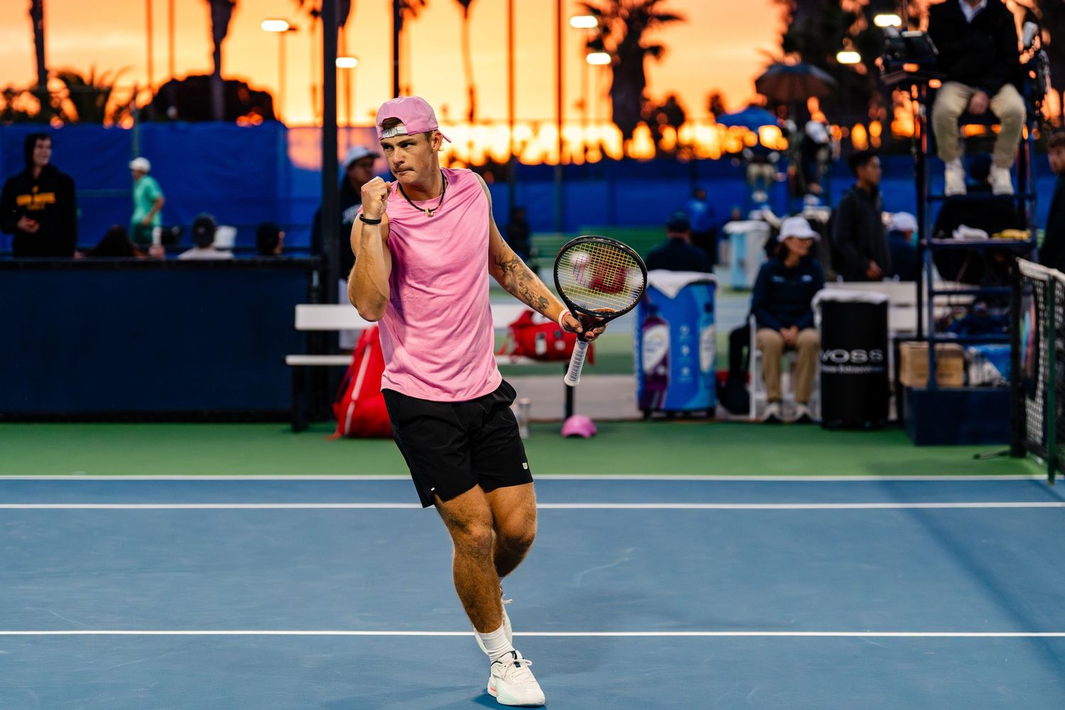 Tennis player in pink shirt celebrates on court at sunset during the San Diego Open tournament.