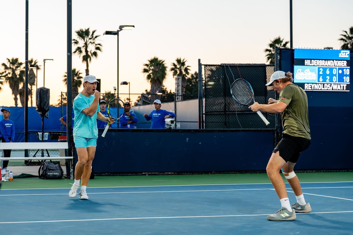 Two male tennis players practice doubles on a blue hard court at dusk with palm trees in background.