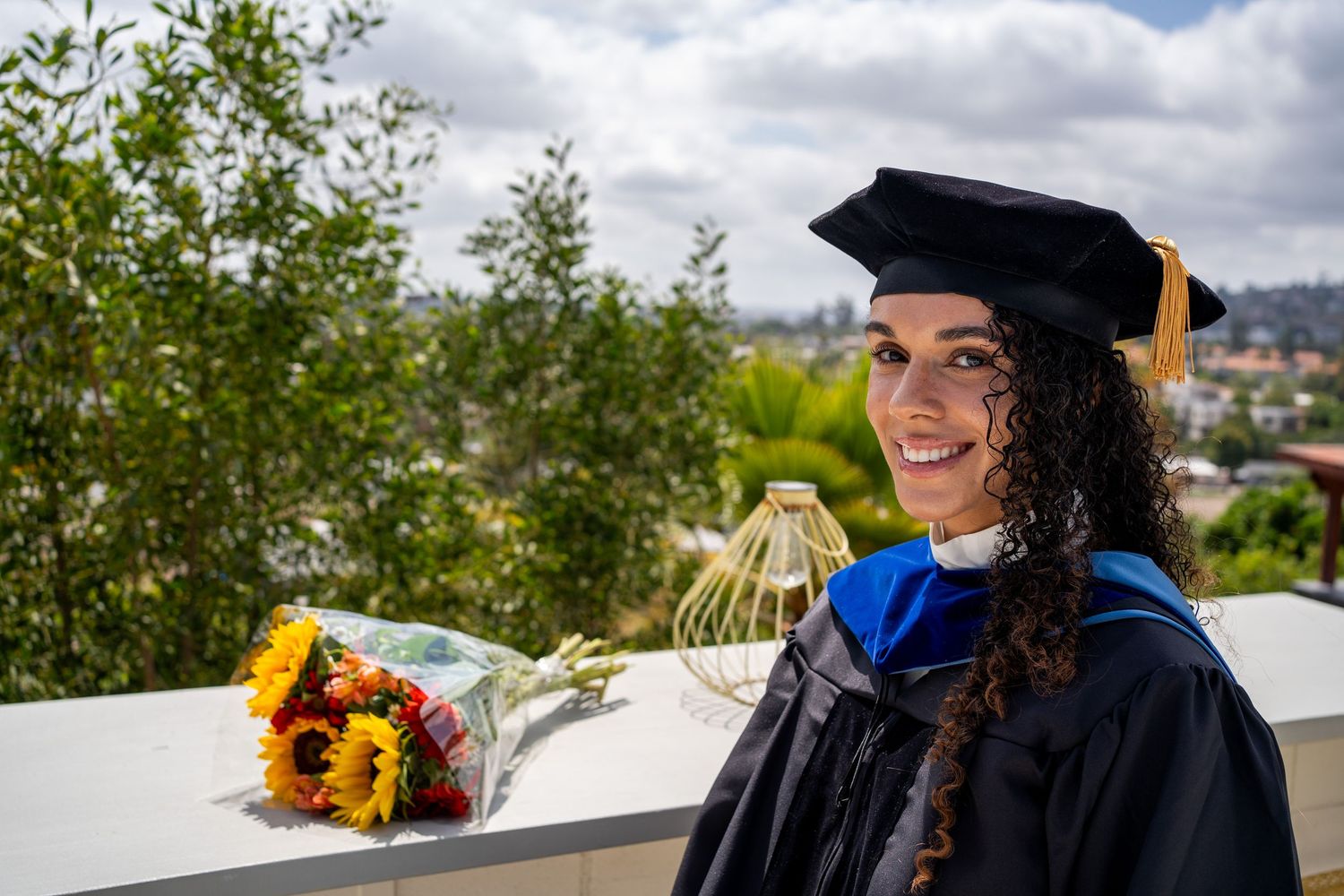 Happy graduate in cap and gown holding sunflower bouquet outdoors with scenic view behind her.