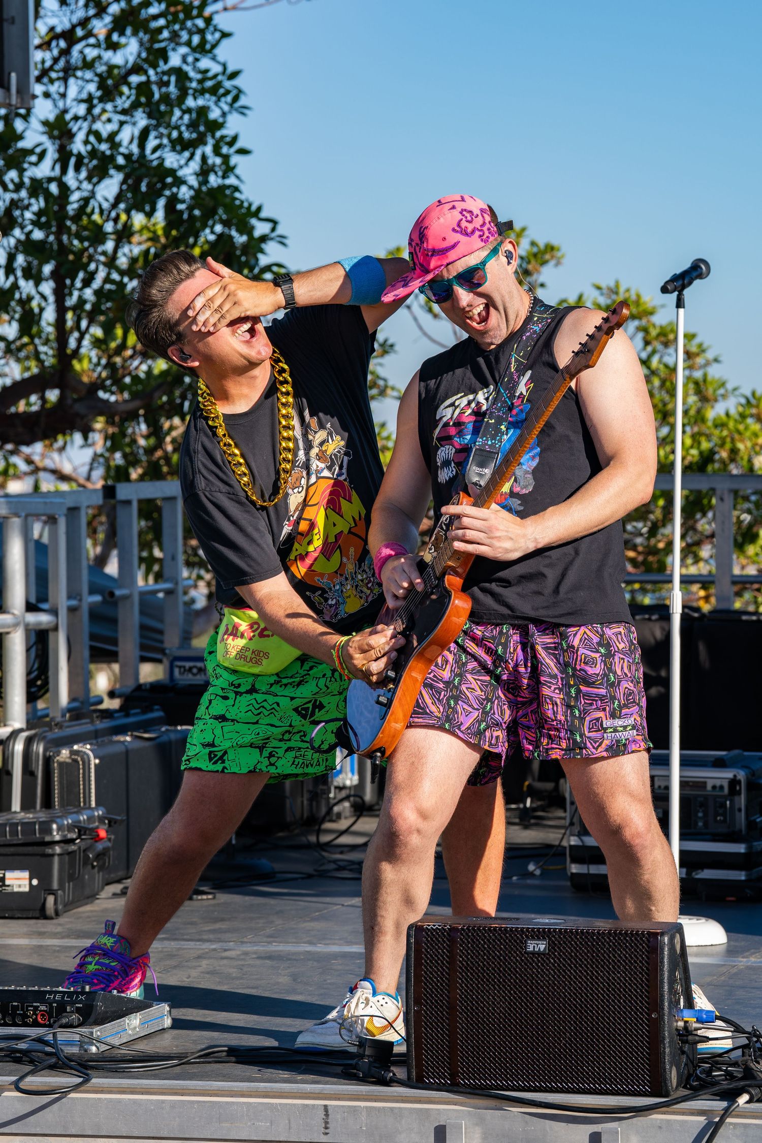 Two colorful musicians perform on an outdoor stage, one playing guitar while the other playfully covers their eyes.