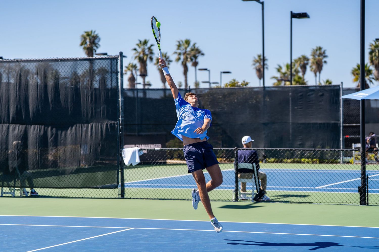 Tennis player leaps to serve on an outdoor court with palm trees behind.