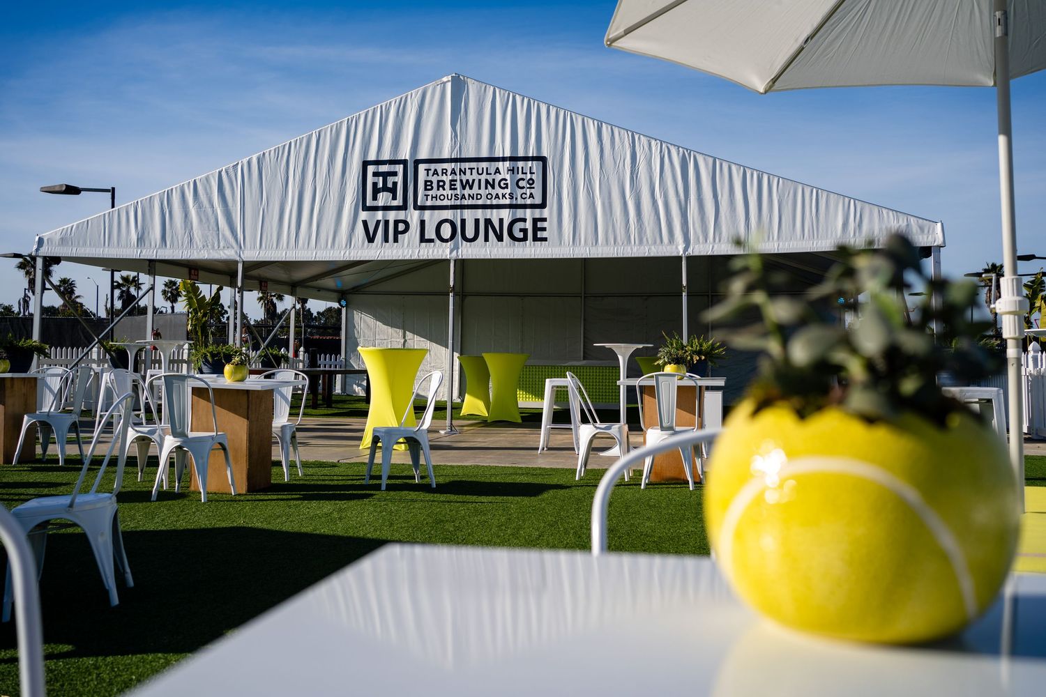 White VIP Lounge tent with yellow chairs and tennis ball decorations at an outdoor tennis tournament.