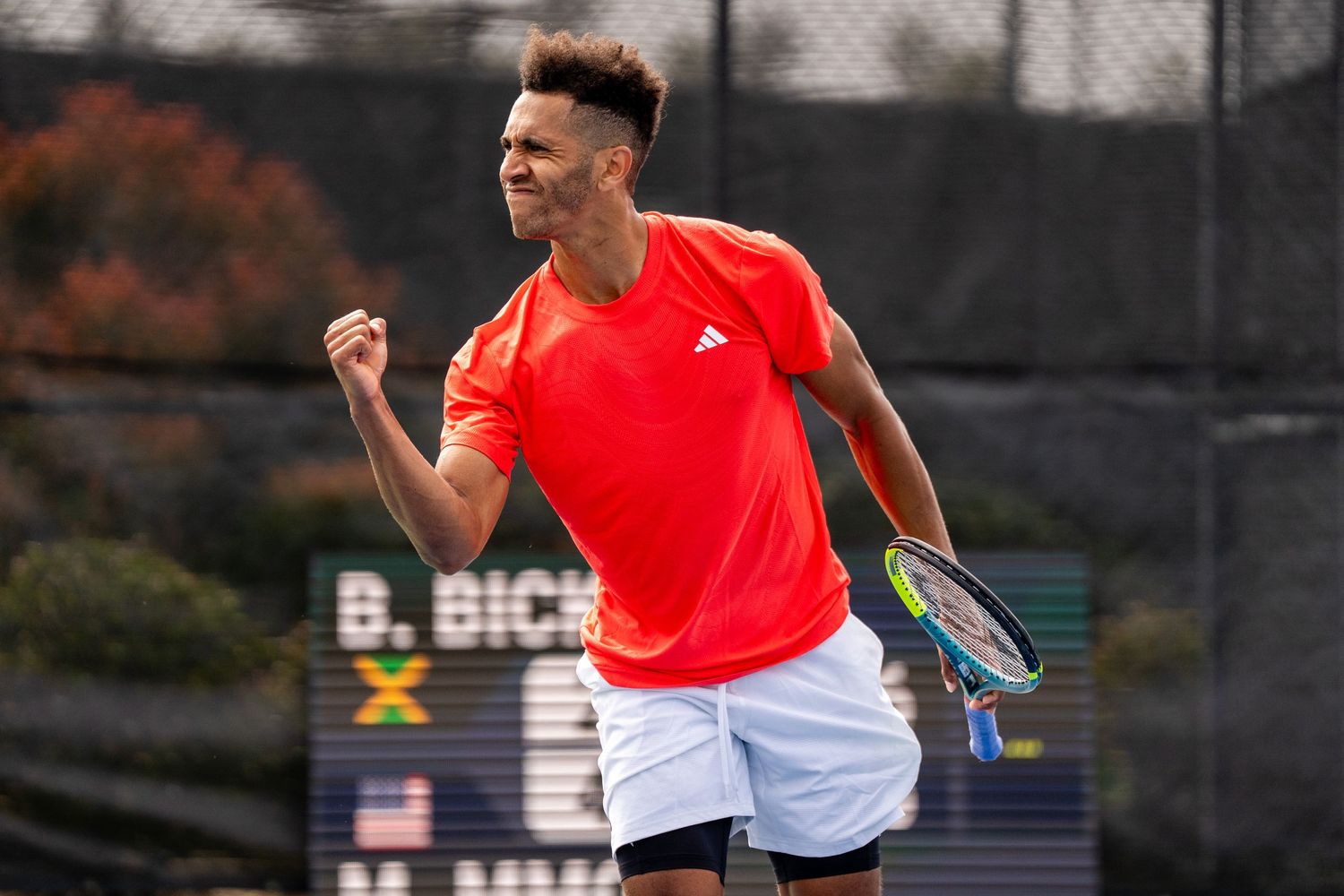 Tennis player in red Adidas shirt celebrates with fist pump on outdoor court.