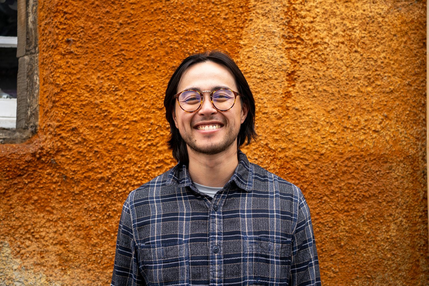 Smiling young man with glasses and plaid shirt poses against textured orange wall outdoors.
