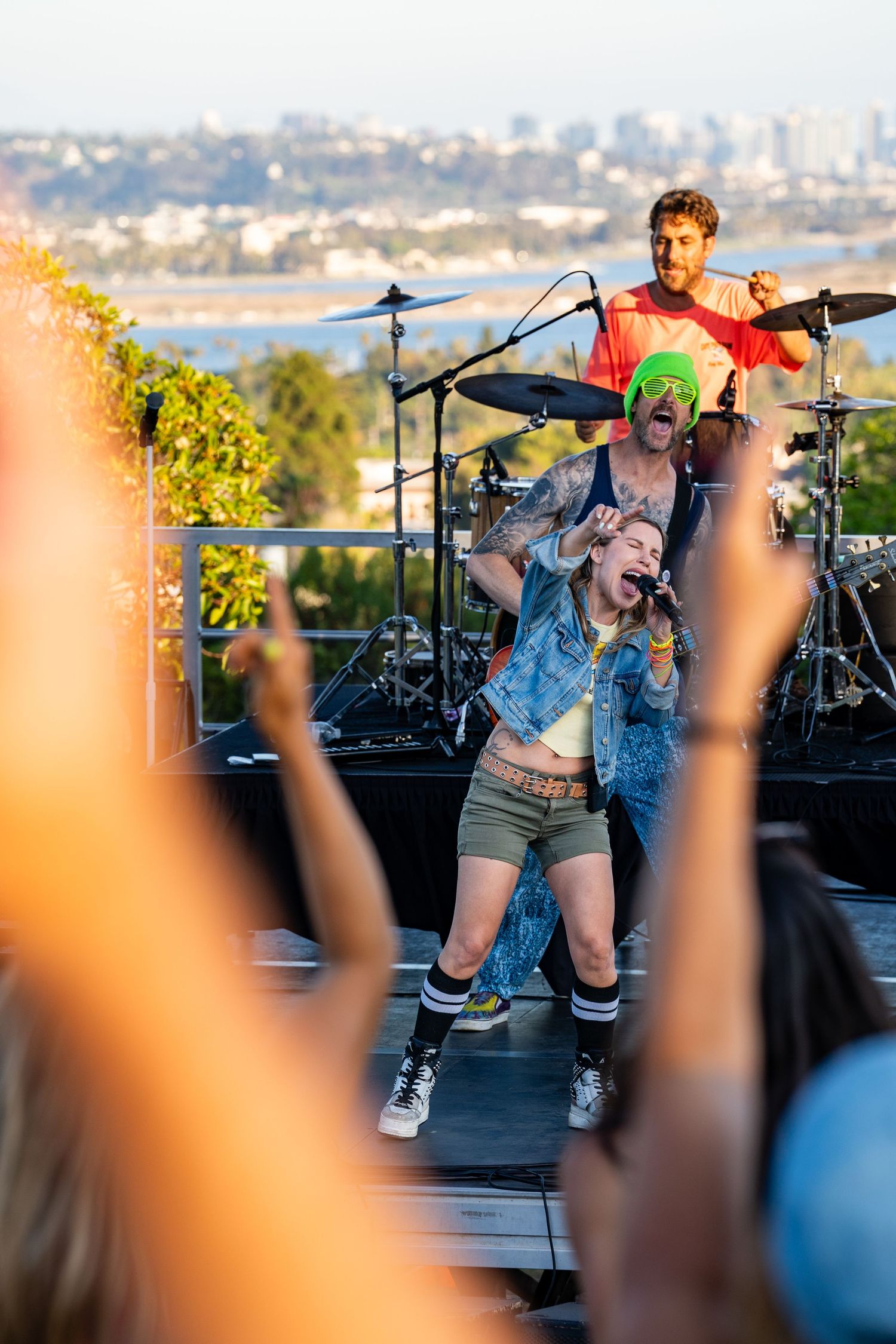 Dynamic live band performance with female singer and drummer on an outdoor stage overlooking a scenic cityscape.