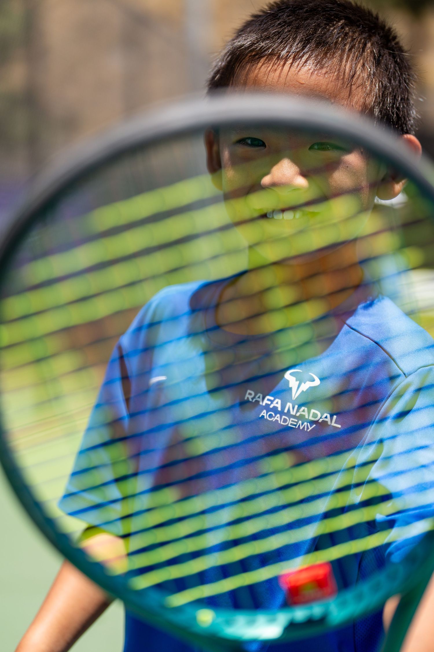 Young boy at Rafa Nadal Academy holding a tennis racket up to the camera on a sunny court.