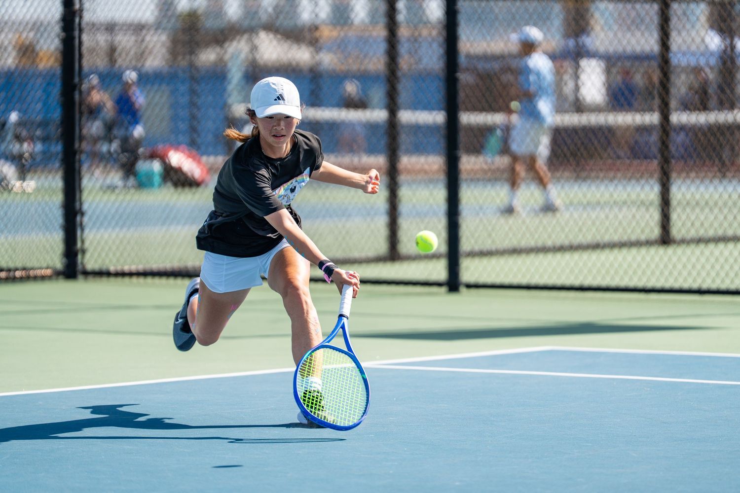 Tennis player in black lunging to hit a low forehand shot on an outdoor blue court.