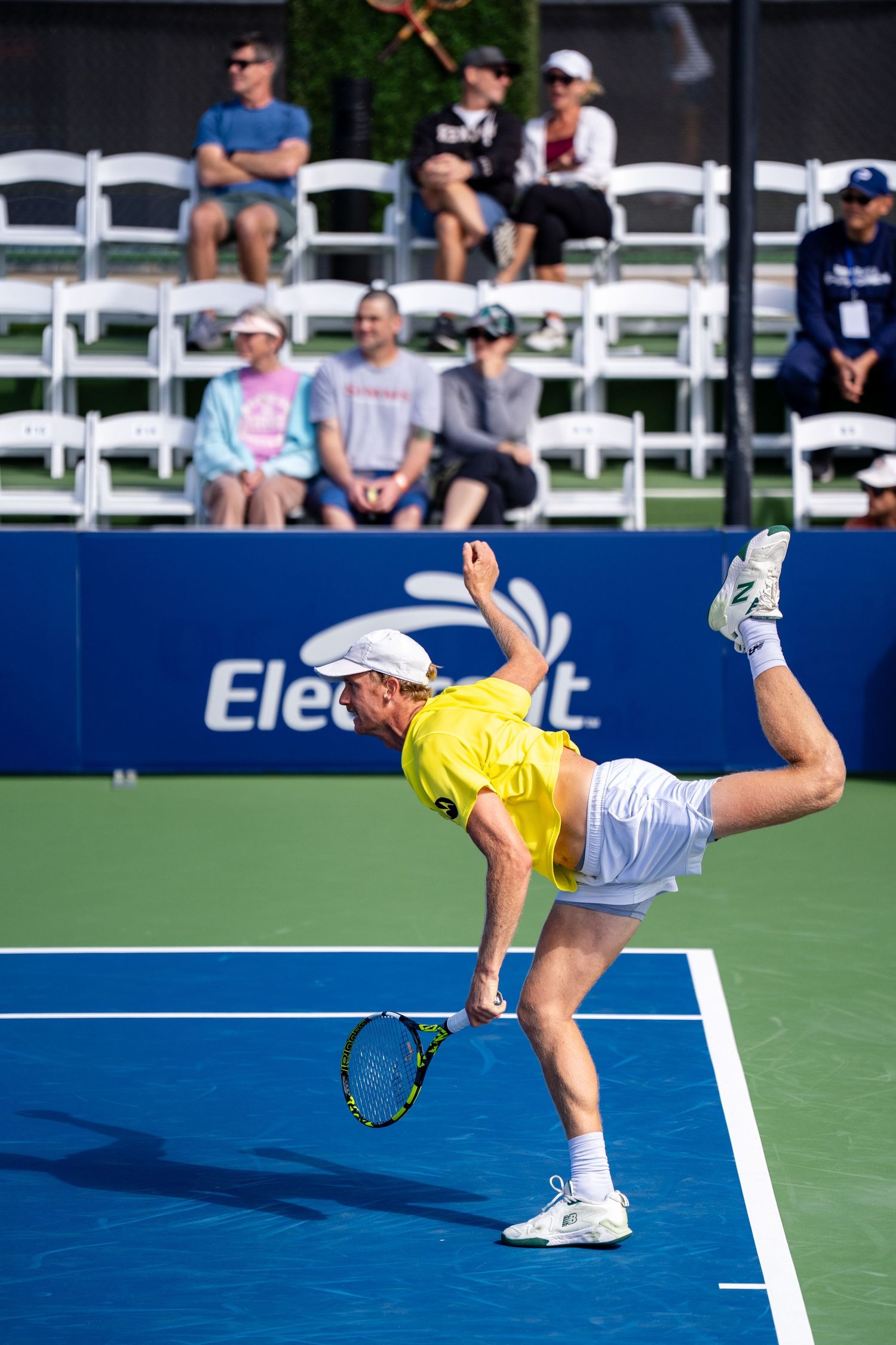 Tennis player in yellow shirt serving with high leg kick on blue outdoor court.