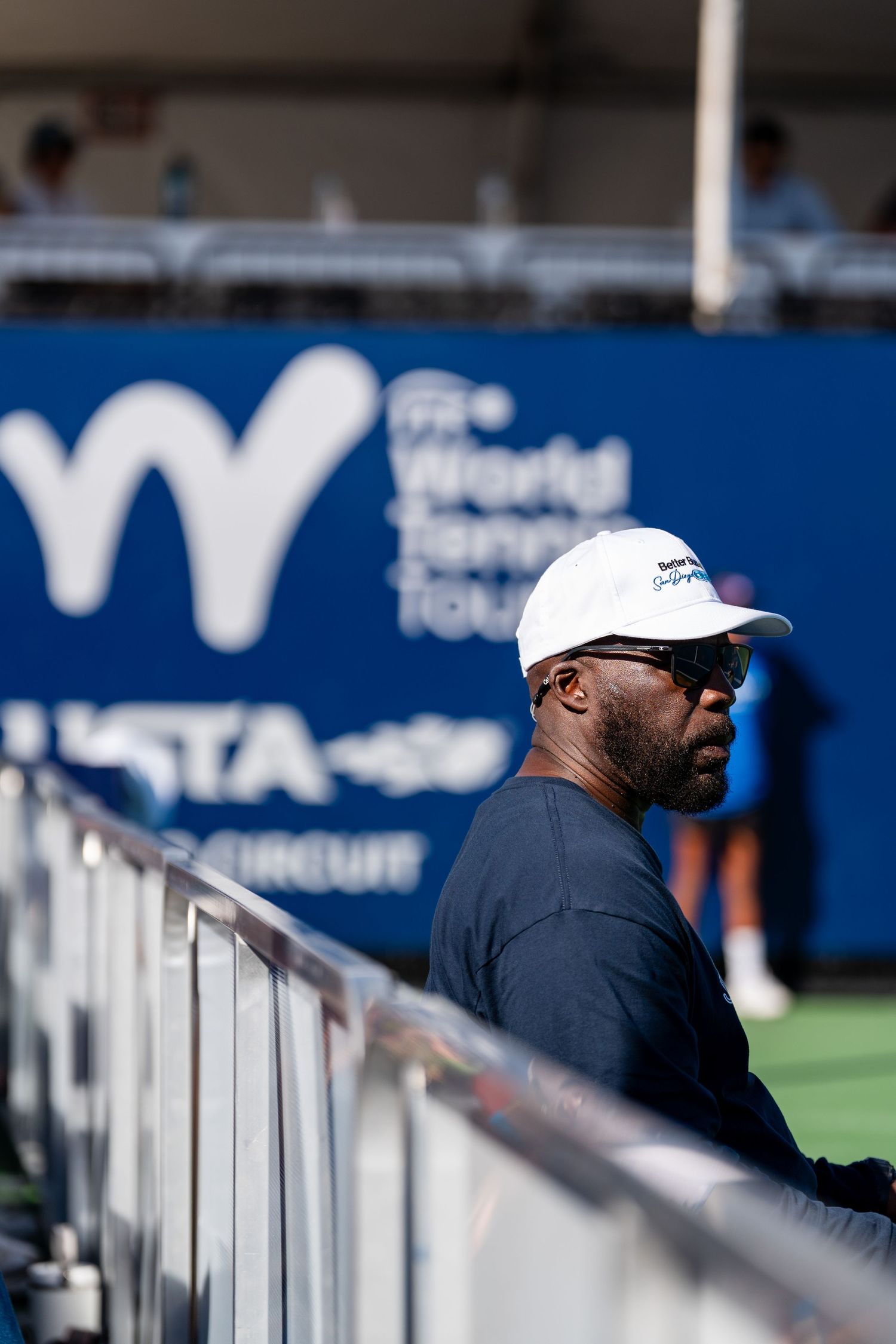 Man in white cap and navy shirt watching a tennis match at the ITF World Tennis Tour USTA Pro Circuit event.