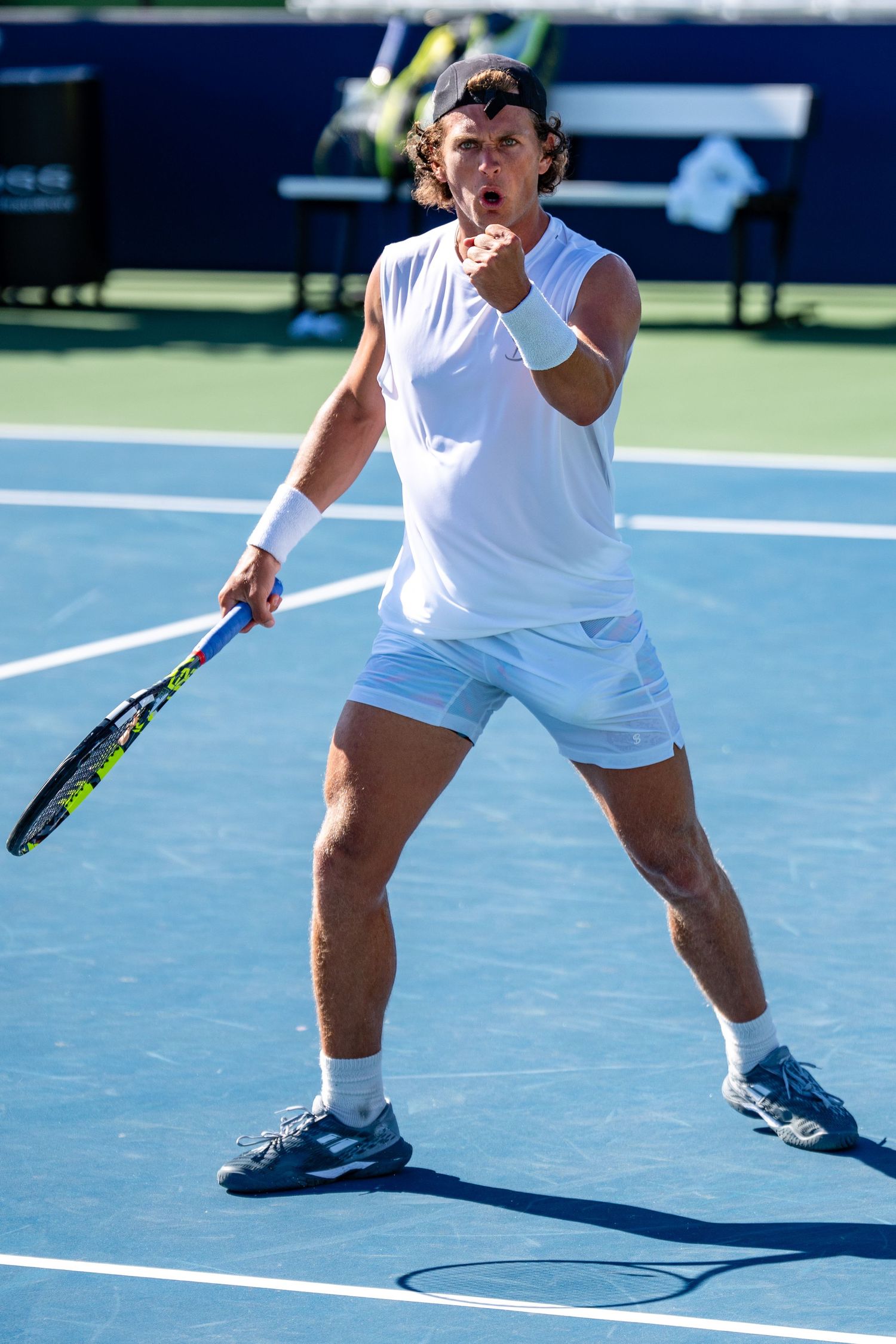 Male tennis player in white outfit celebrating a point with fist pump on blue hard court.