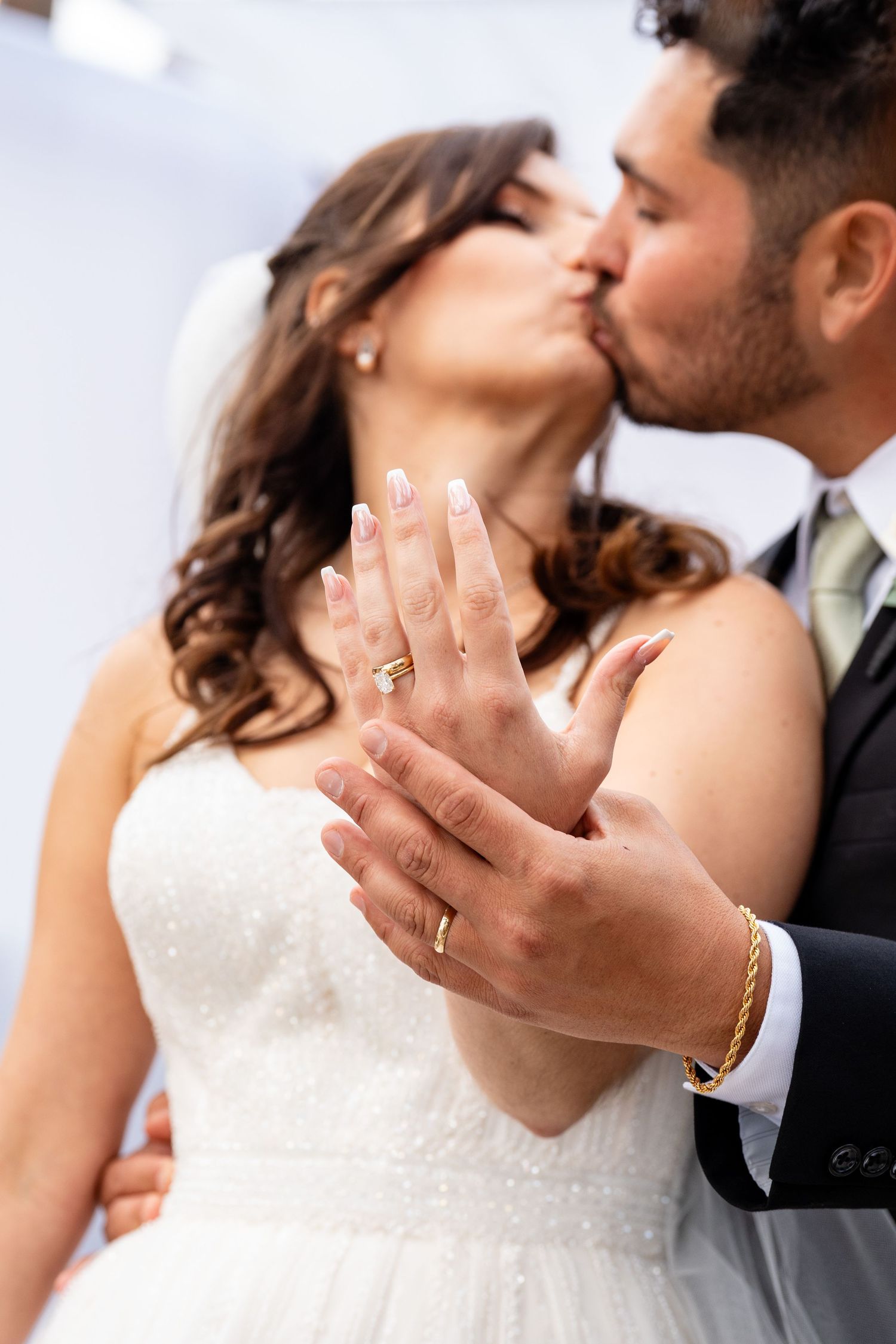 Newlywed couple kissing while showing off their gold wedding rings, bride in white gown with veil, groom in dark suit.