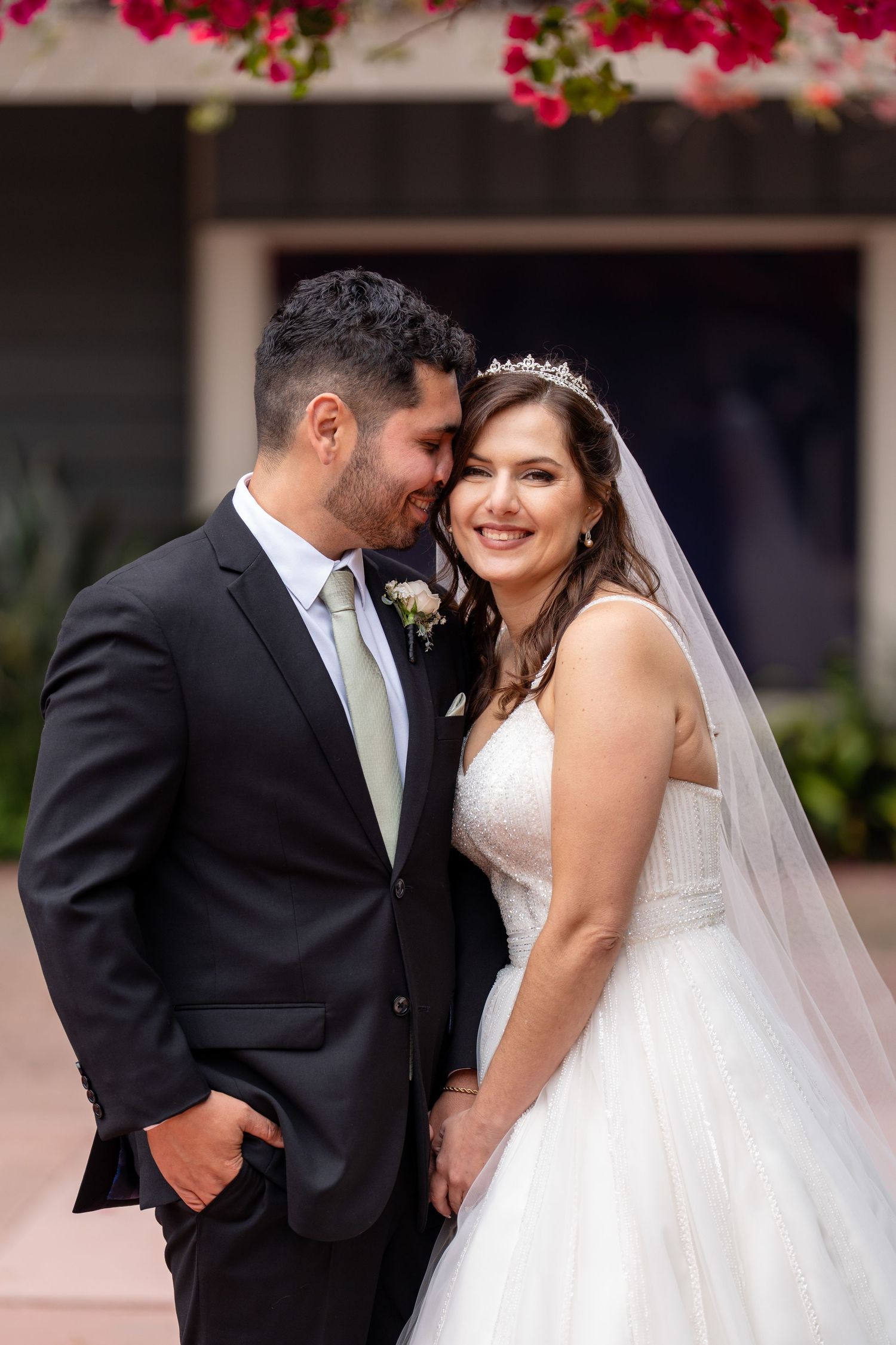 A bride in a white gown and tiara smiles as her groom in a dark suit kisses her cheek outdoors.