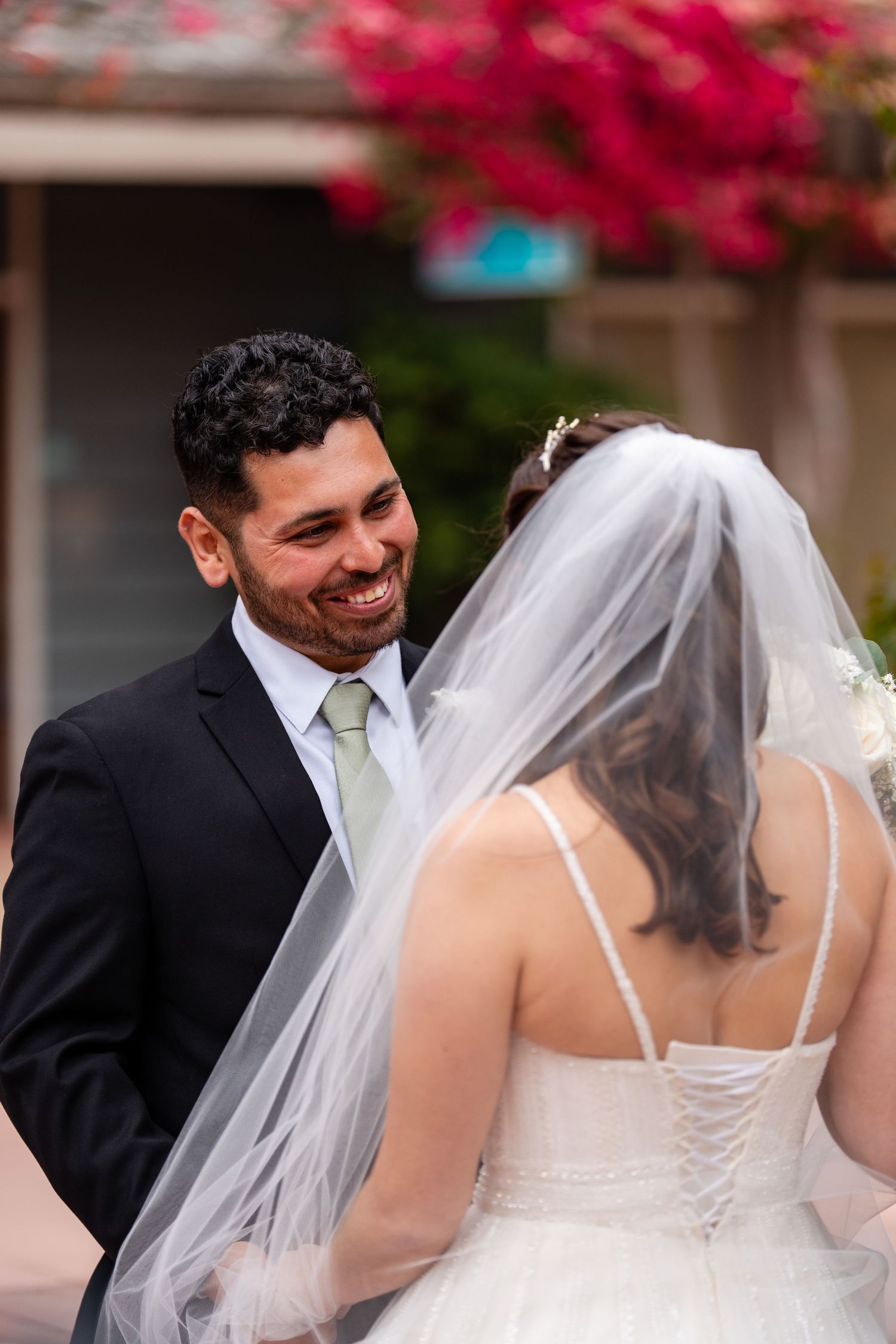 Smiling groom in black suit with green tie facing bride in white wedding dress and veil outdoors.