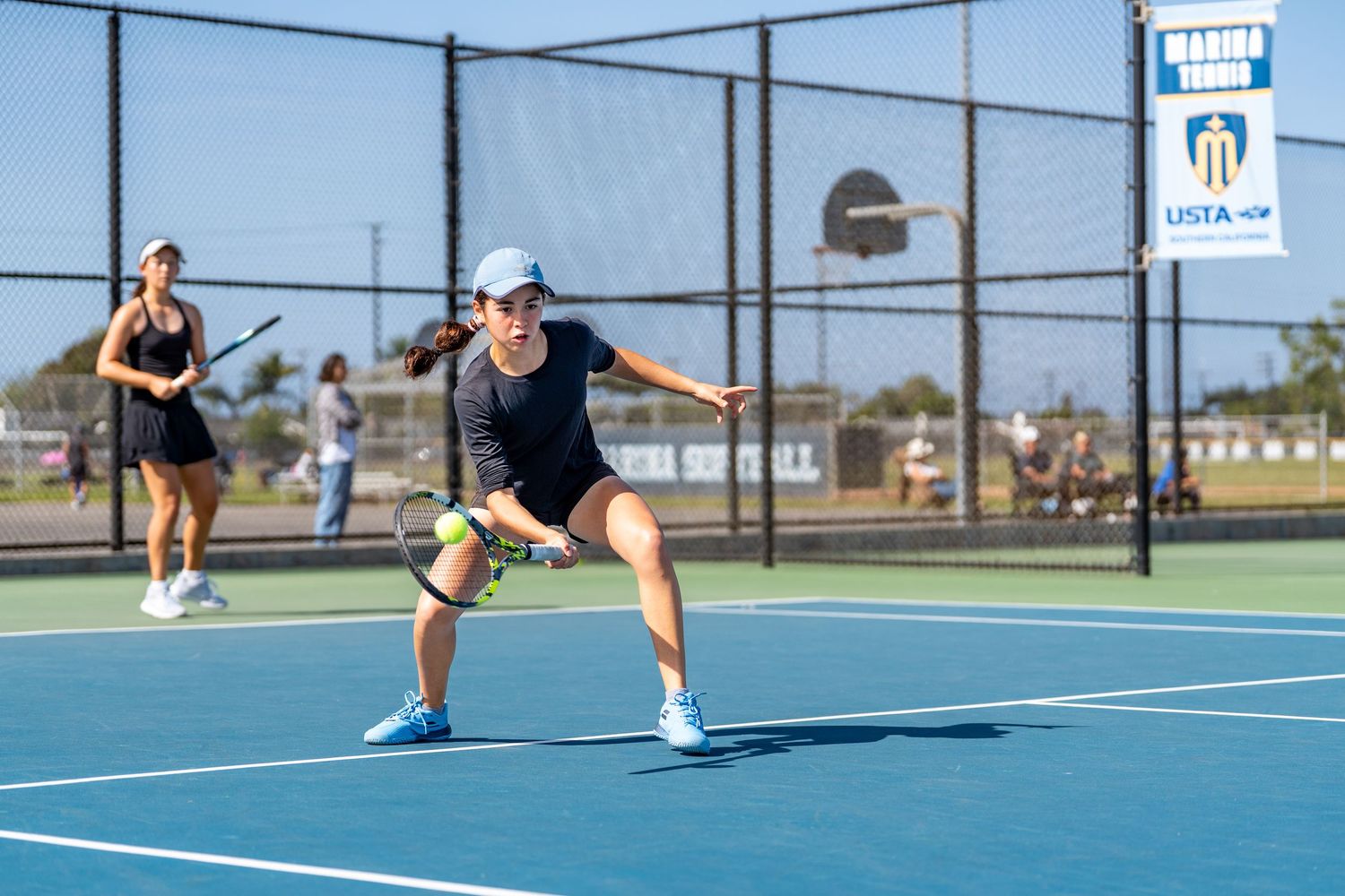 Female tennis player in black outfit preparing to return a shot on an outdoor tennis court.