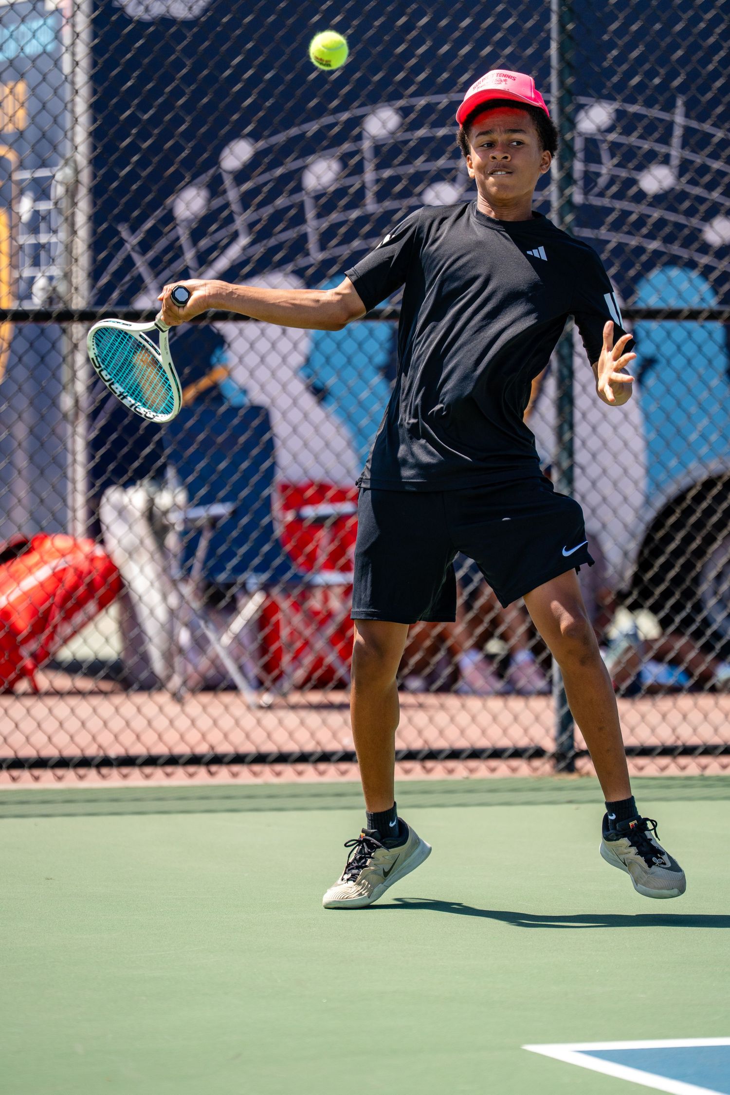 Young male tennis player in black and red hits a forehand shot on an outdoor court.