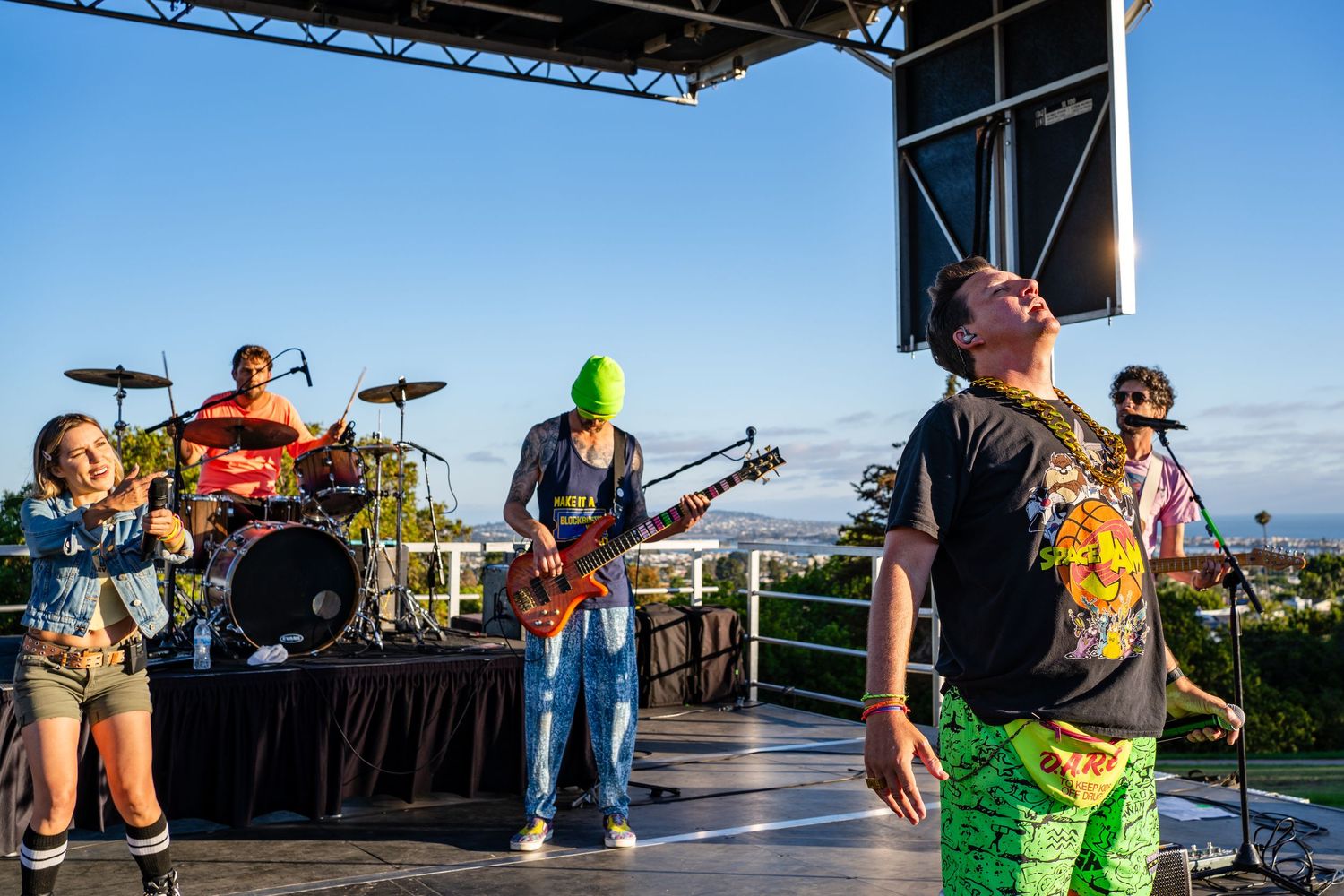 Colorful band performing on outdoor stage at sunset with guitarist in green mask and vibrant costumes.