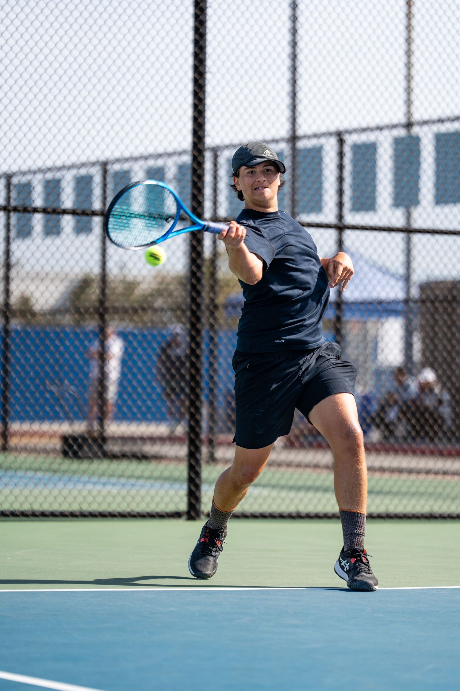 Male tennis player in black outfit hitting a forehand shot on an outdoor blue tennis court.