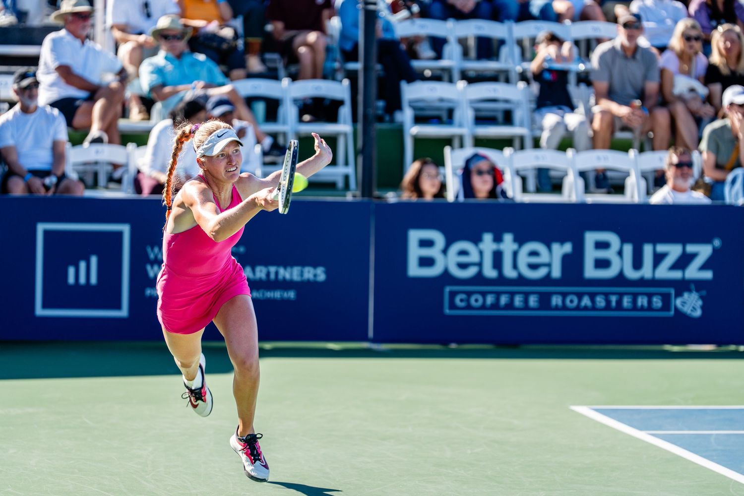 Female tennis player in pink dress hitting a forehand shot before a crowd at the Better Buzz Coffee Roasters tennis event.