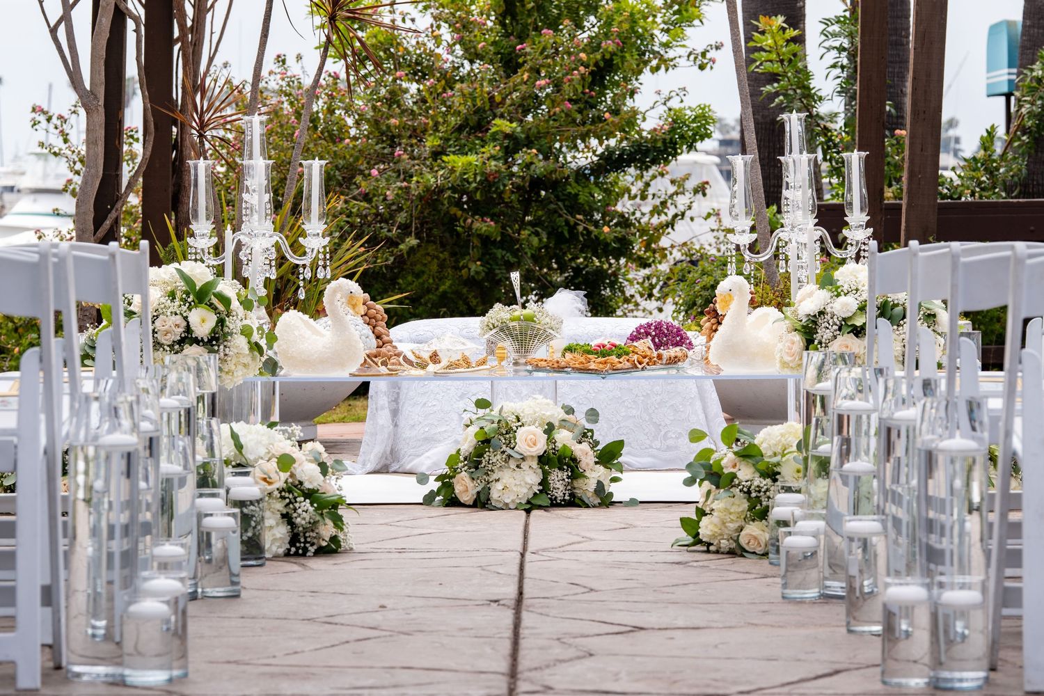 Elegant outdoor wedding ceremony aisle with white floral arrangements, candles, and a decorated altar table.