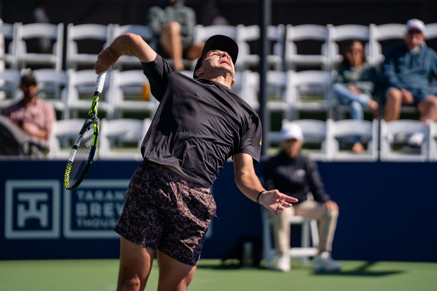 Tennis player in black outfit serves powerfully during an outdoor professional tennis tournament match.