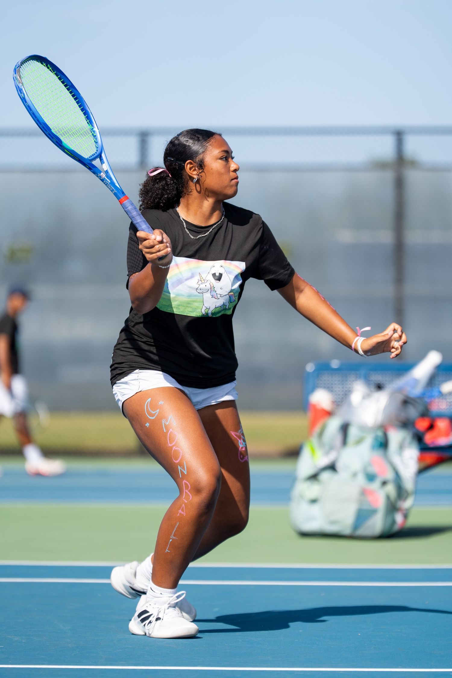 Female tennis player in black graphic tee executing a forehand stroke on a sunny outdoor court.