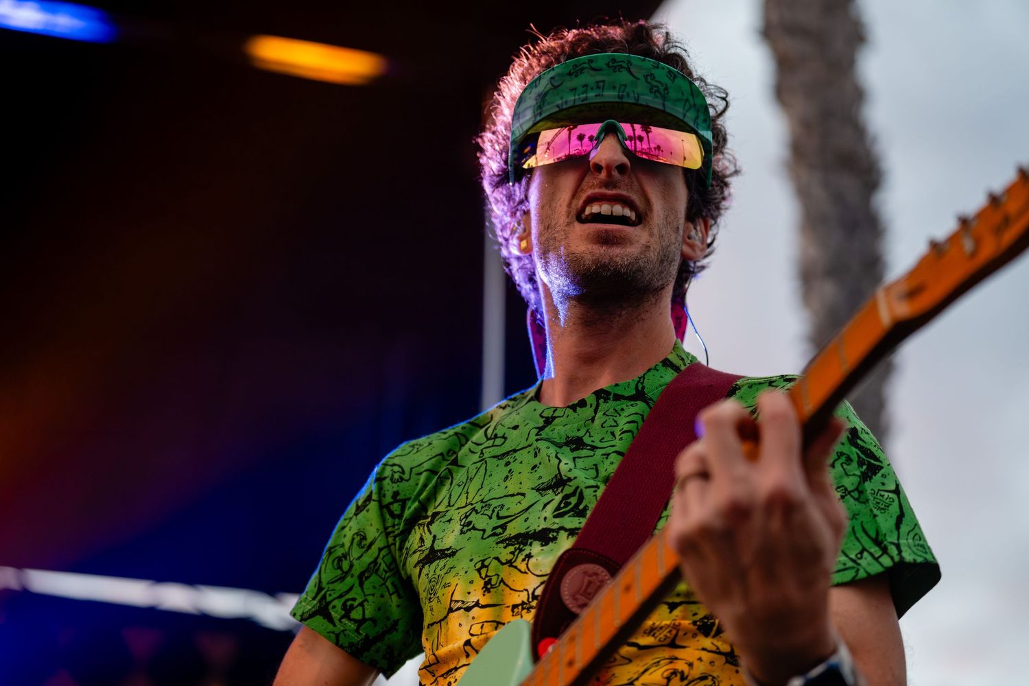 Smiling guitarist wearing pink sunglasses and green patterned shirt performing on stage outdoors.