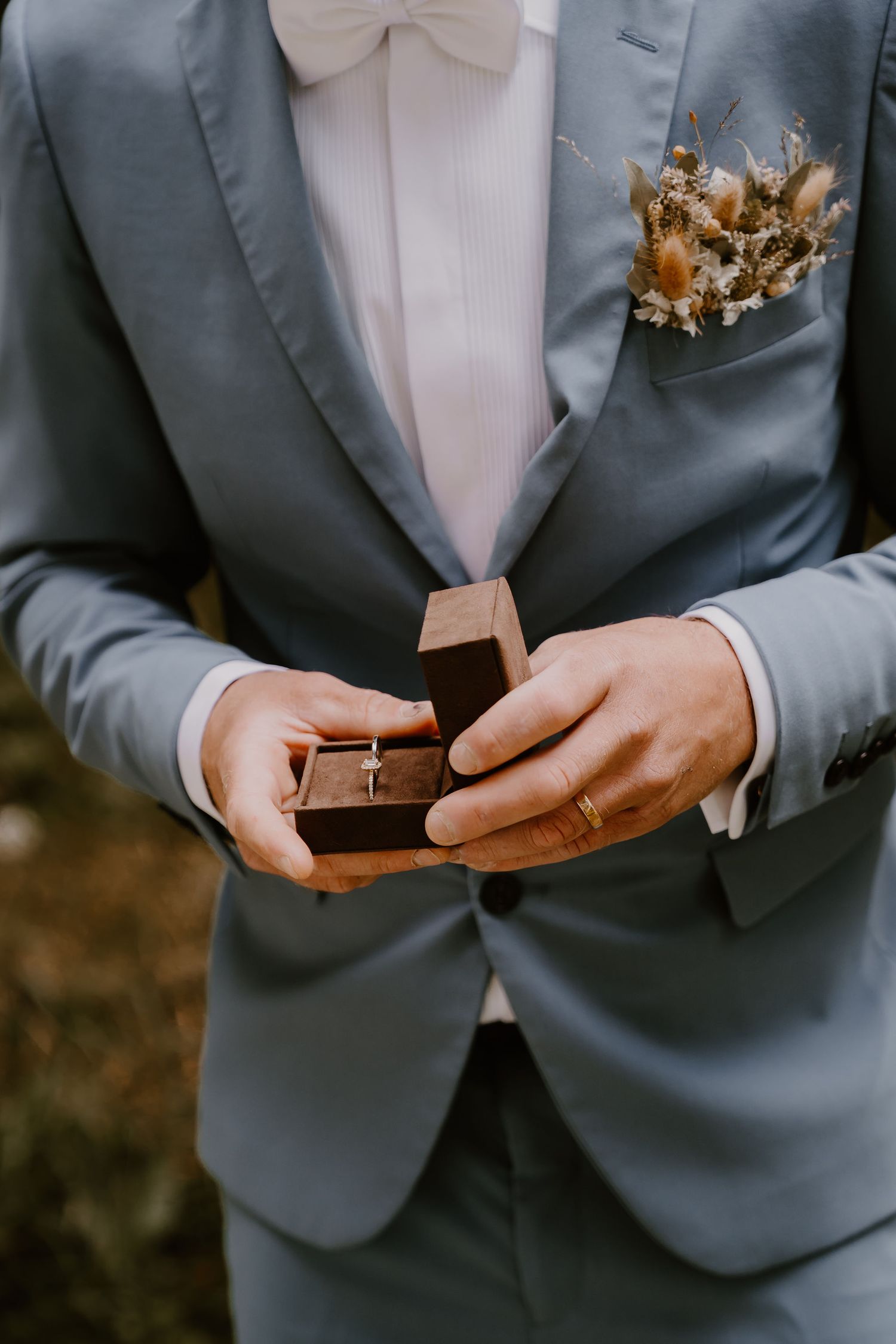 A person in a grey suit and bow tie holds and opens a simple wooden ring box.