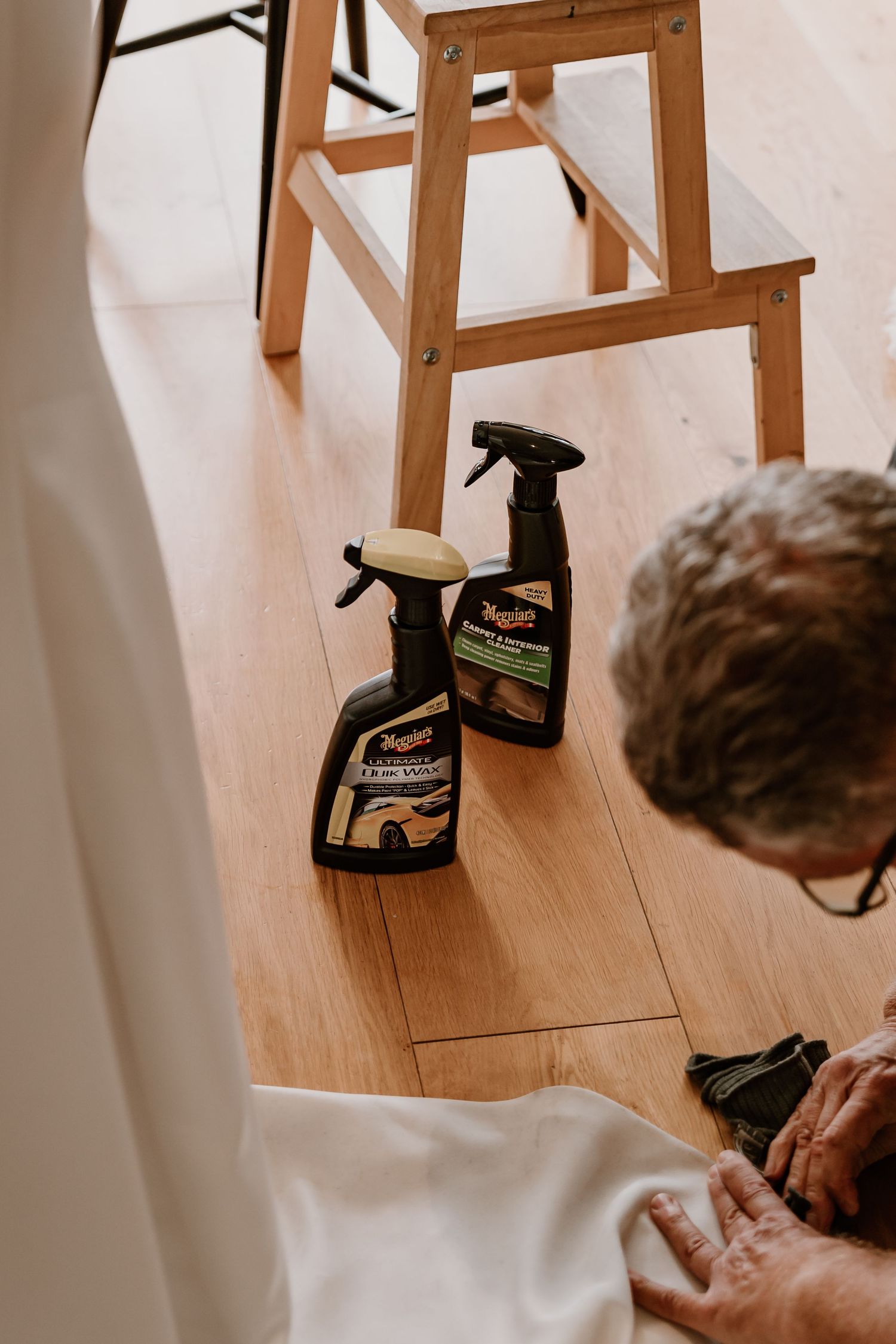 Someone holds a cleaning spray bottle while kneeling on a wood floor near a white wall.