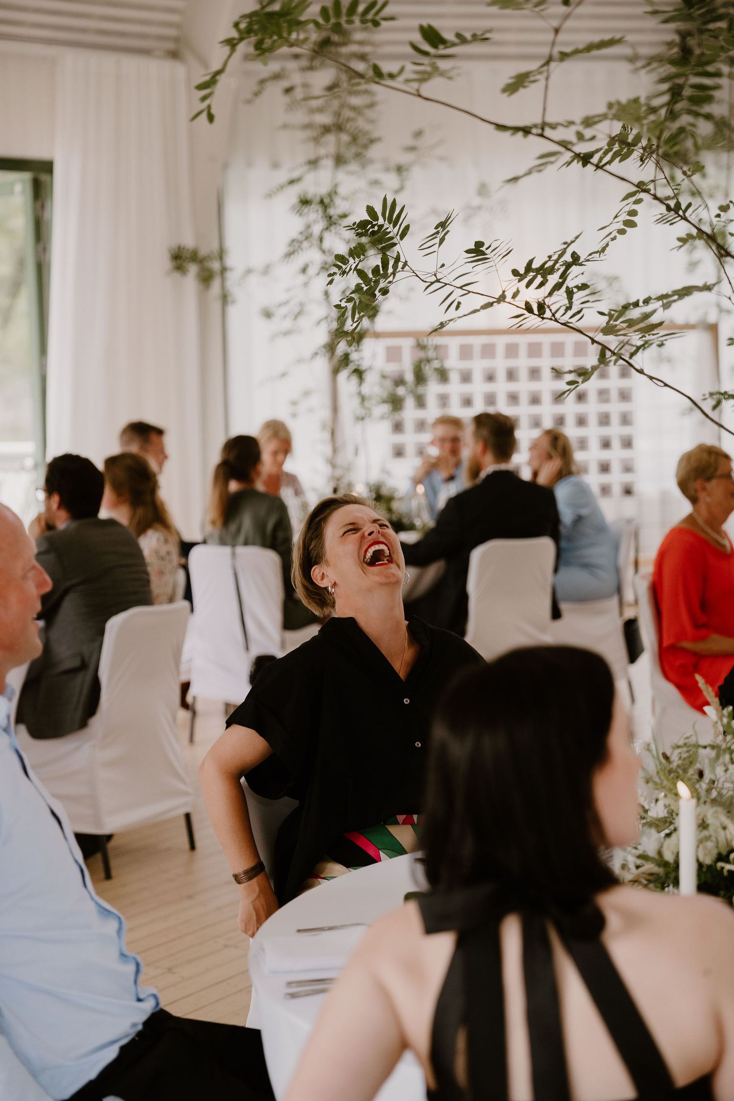 Wedding reception guests mingle and dance in an elegant white-decorated indoor venue with greenery accents.