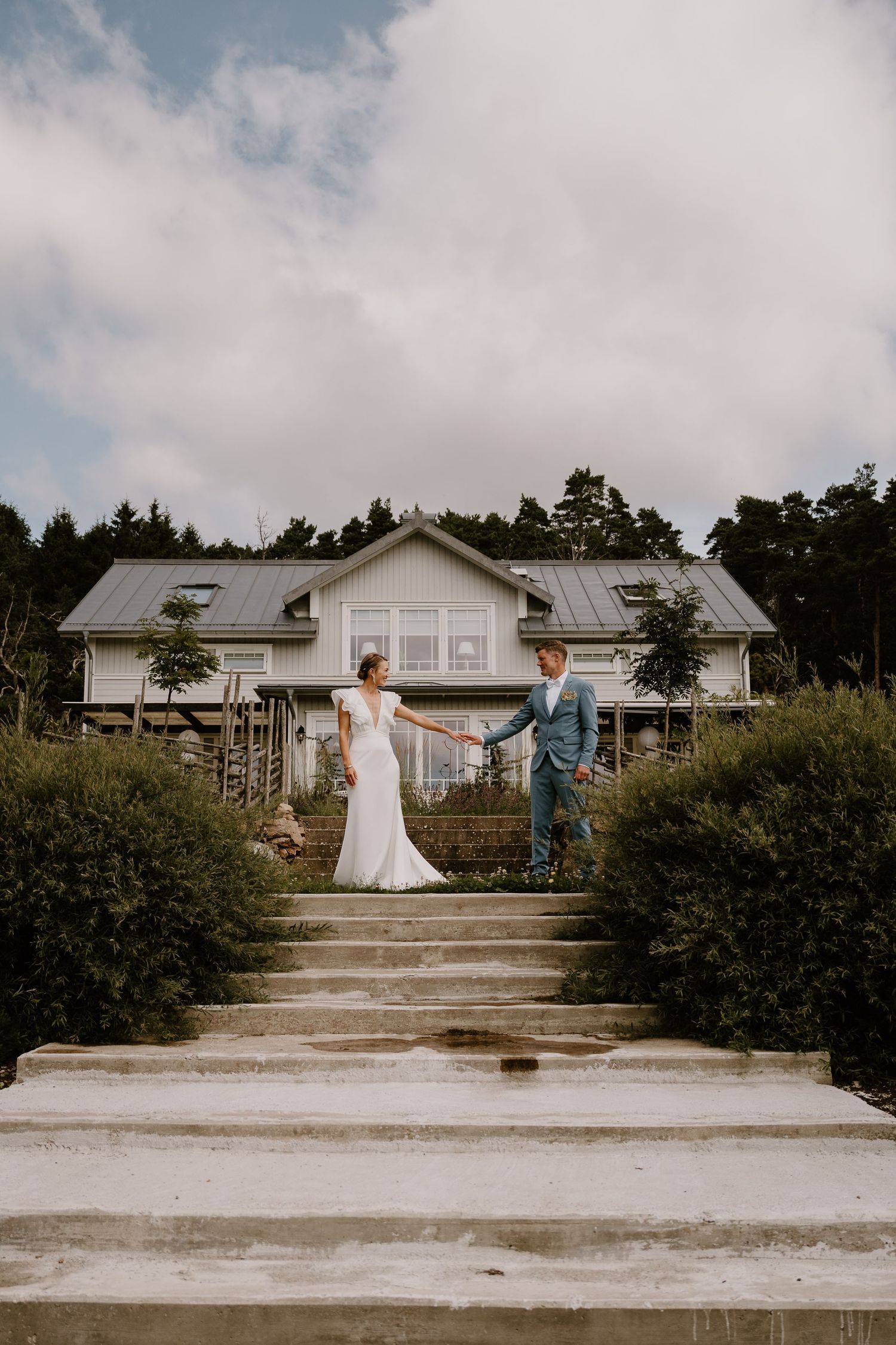 A couple walks on concrete steps leading up to a white house with rustic trees and cloudy skies in the background.