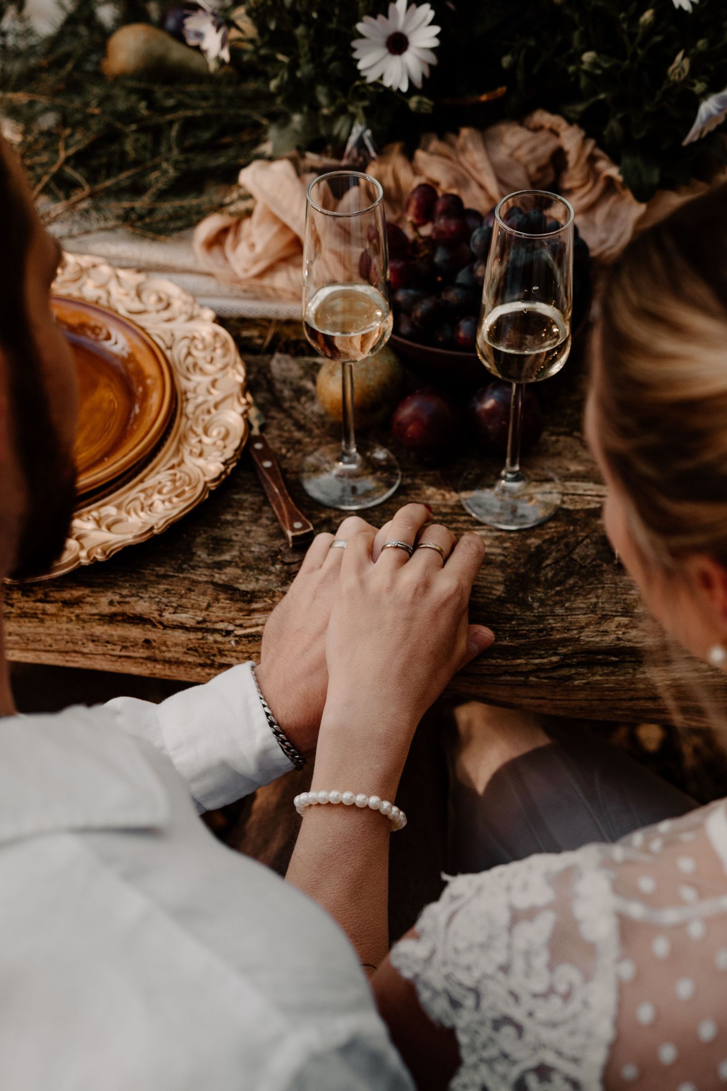 A romantic scene with two wine glasses, a pearl bracelet, and lace details on a rustic wooden table with flowers.