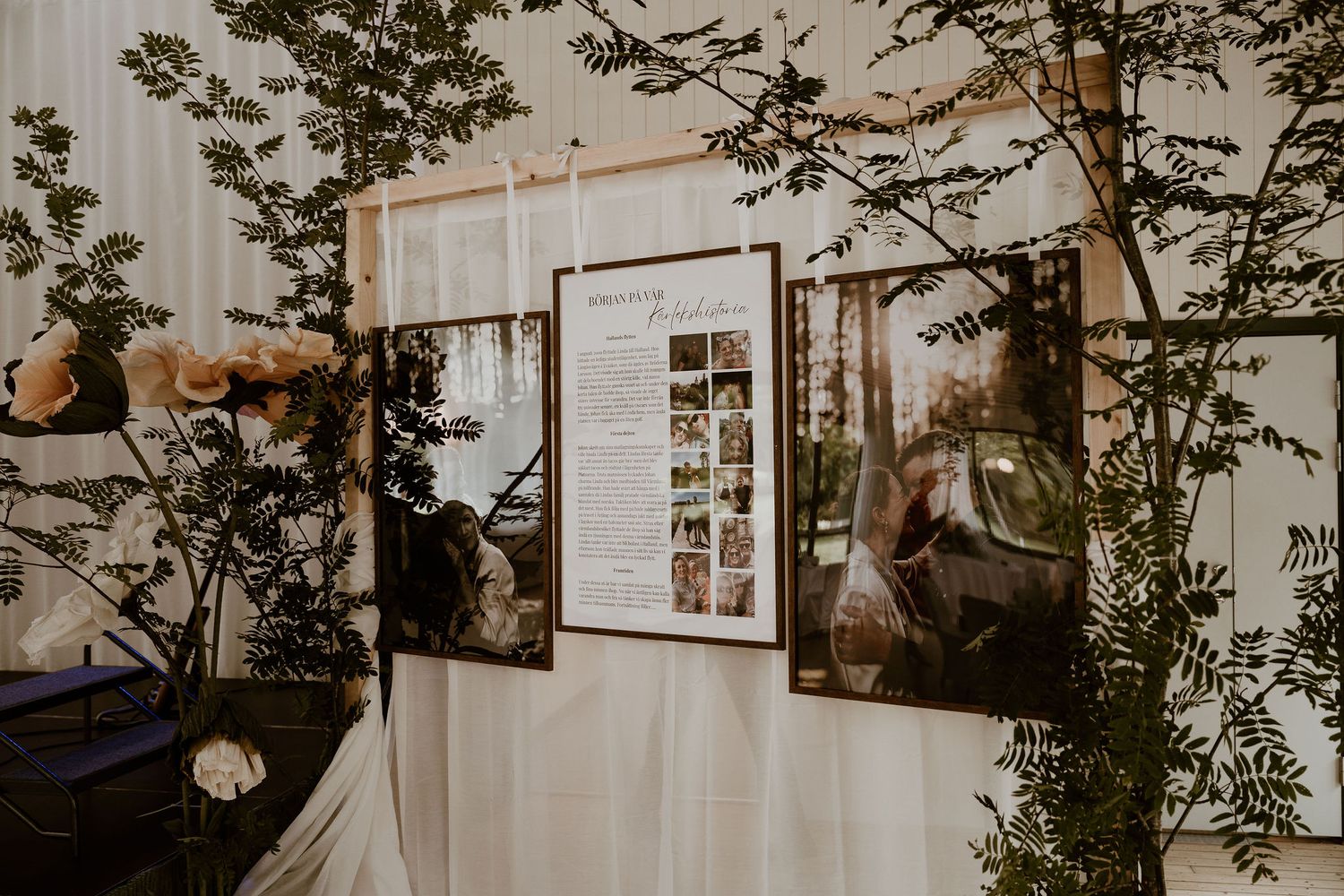A row of white framed gallery photos hang on a wall between tall green indoor plants in warm lighting.