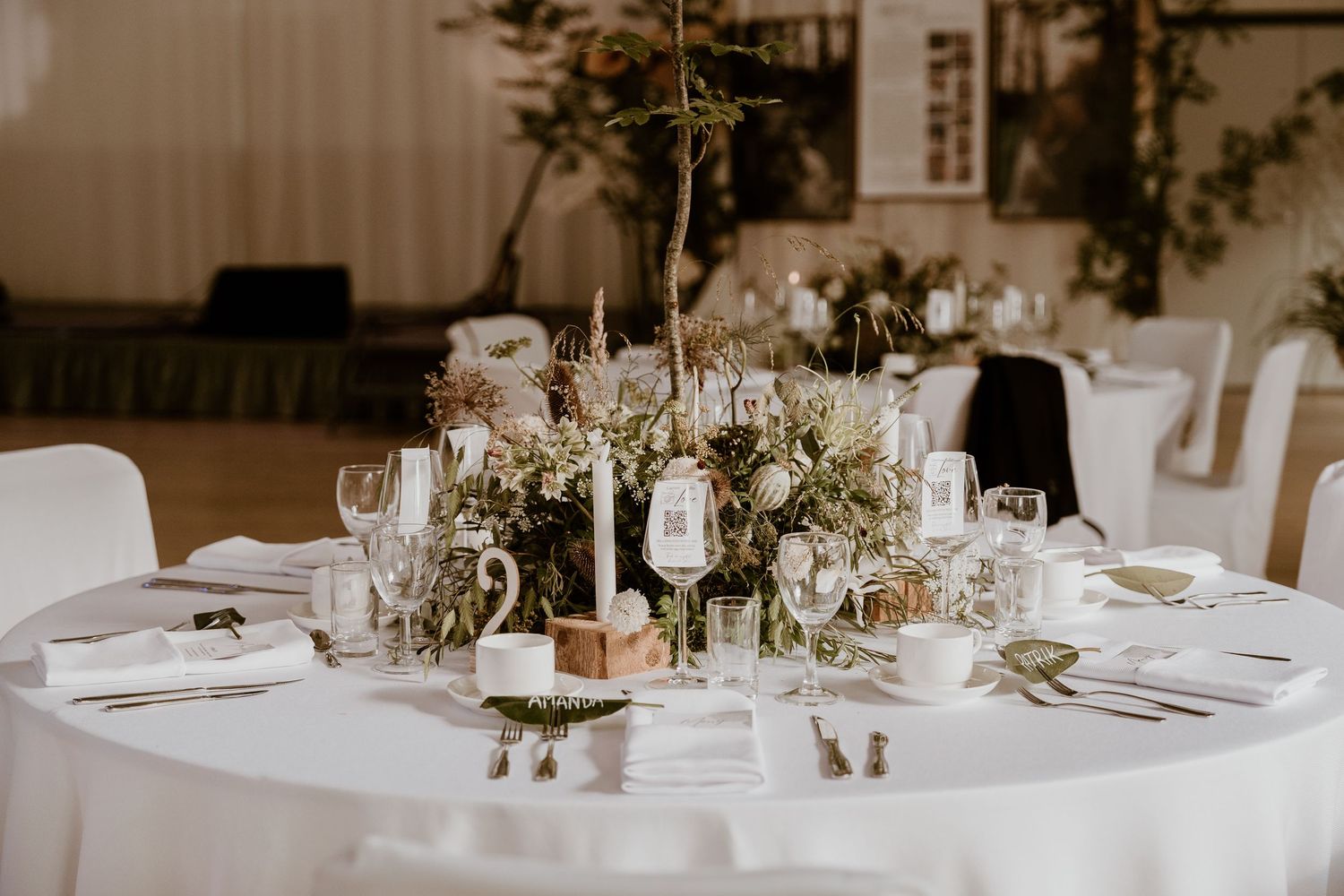 Wedding reception tables decorated with elegant floral centerpieces, white linens, and place settings in a light-filled venue.