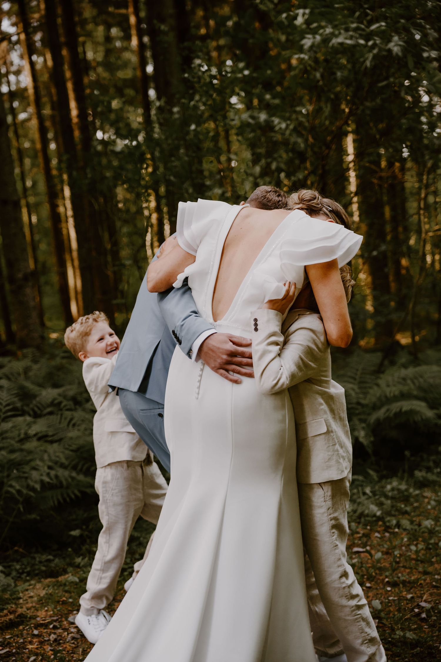 A newlywed couple in formal attire embraces lovingly on a forested trail during their wedding photoshoot.
