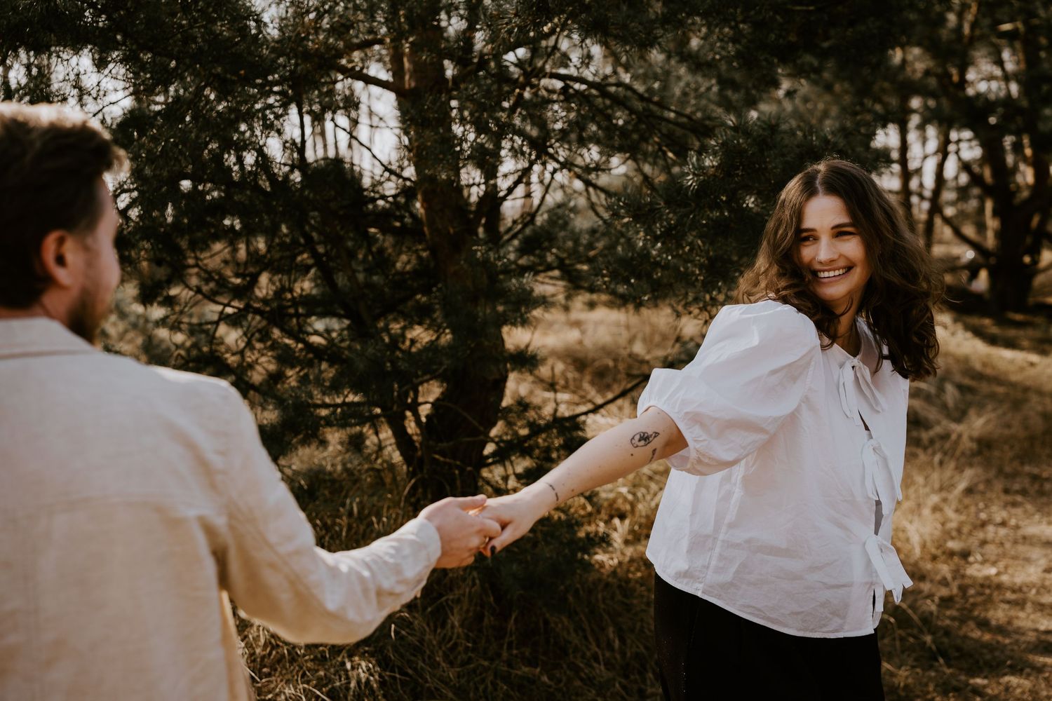 A couple holds hands and smiles while walking through a wooded area at sunset during a romantic outdoor photoshoot.