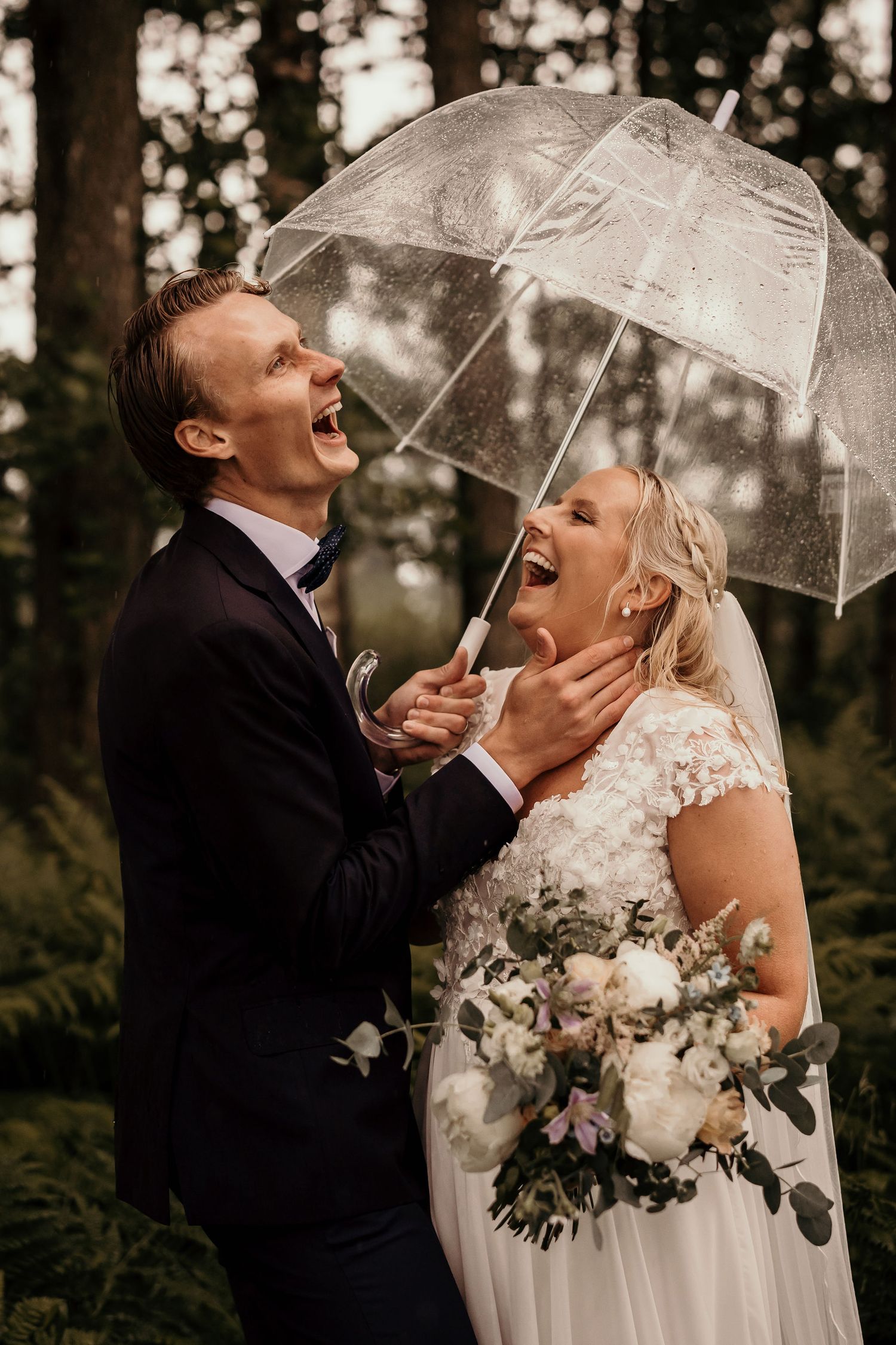 A joyful wedding couple laughs together under a clear umbrella in a forest setting with a beautiful bridal bouquet.