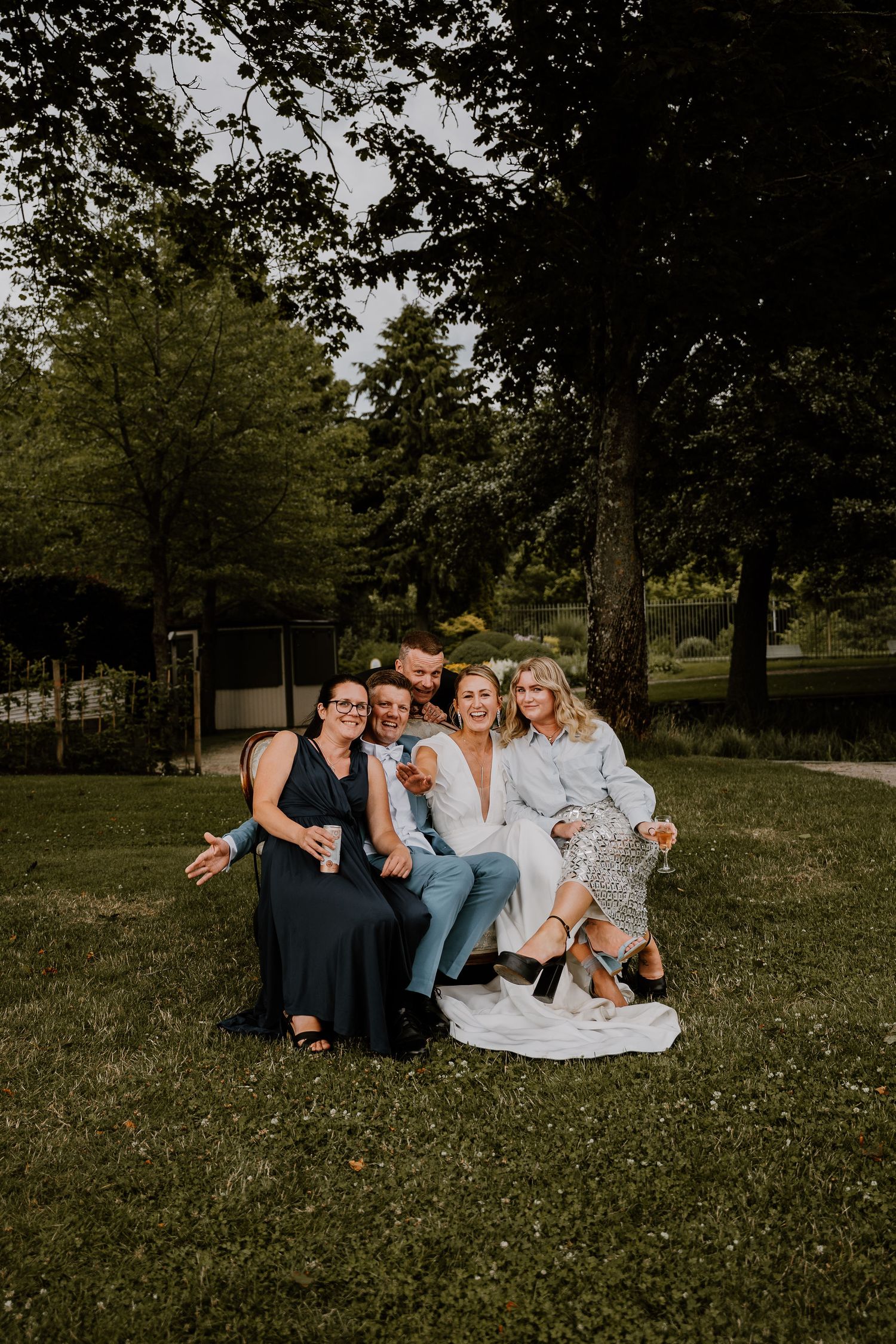 A group sits together on a blanket in a peaceful park setting surrounded by trees on a warm summer day.