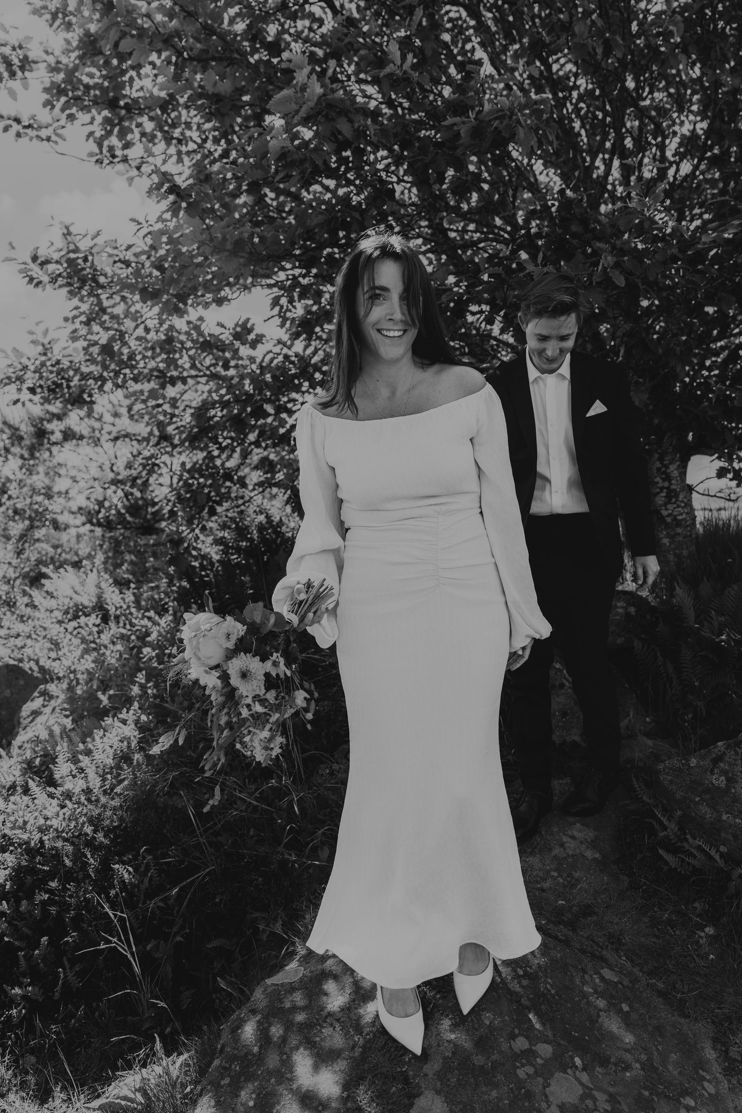 Leende brud i en axelbandslös klänning som håller blommor, går med en brudgum i kostym utomhus