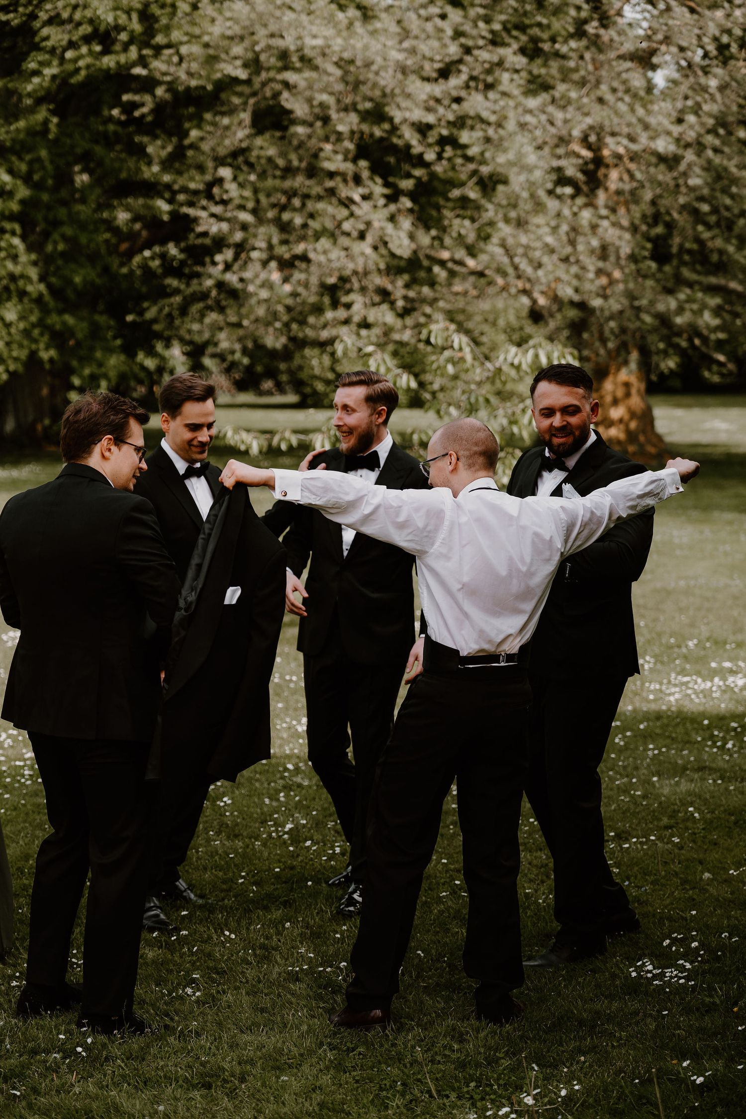 Groomsmen perform a fun domino effect pose falling sideways on a grassy lawn during wedding photos.