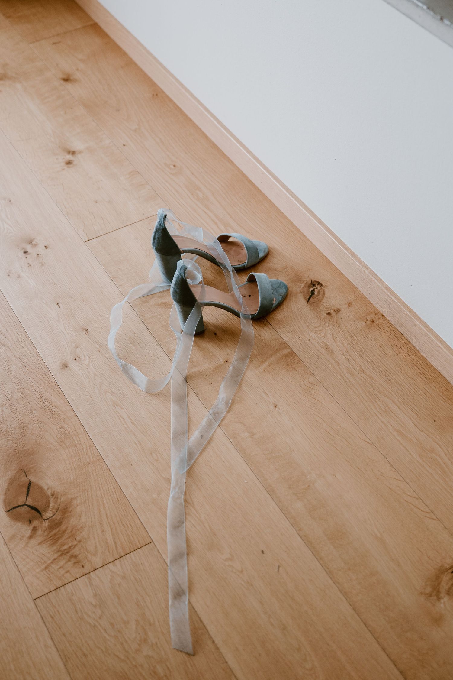A pair of sage green shoes with ribbons sits on light wooden flooring.