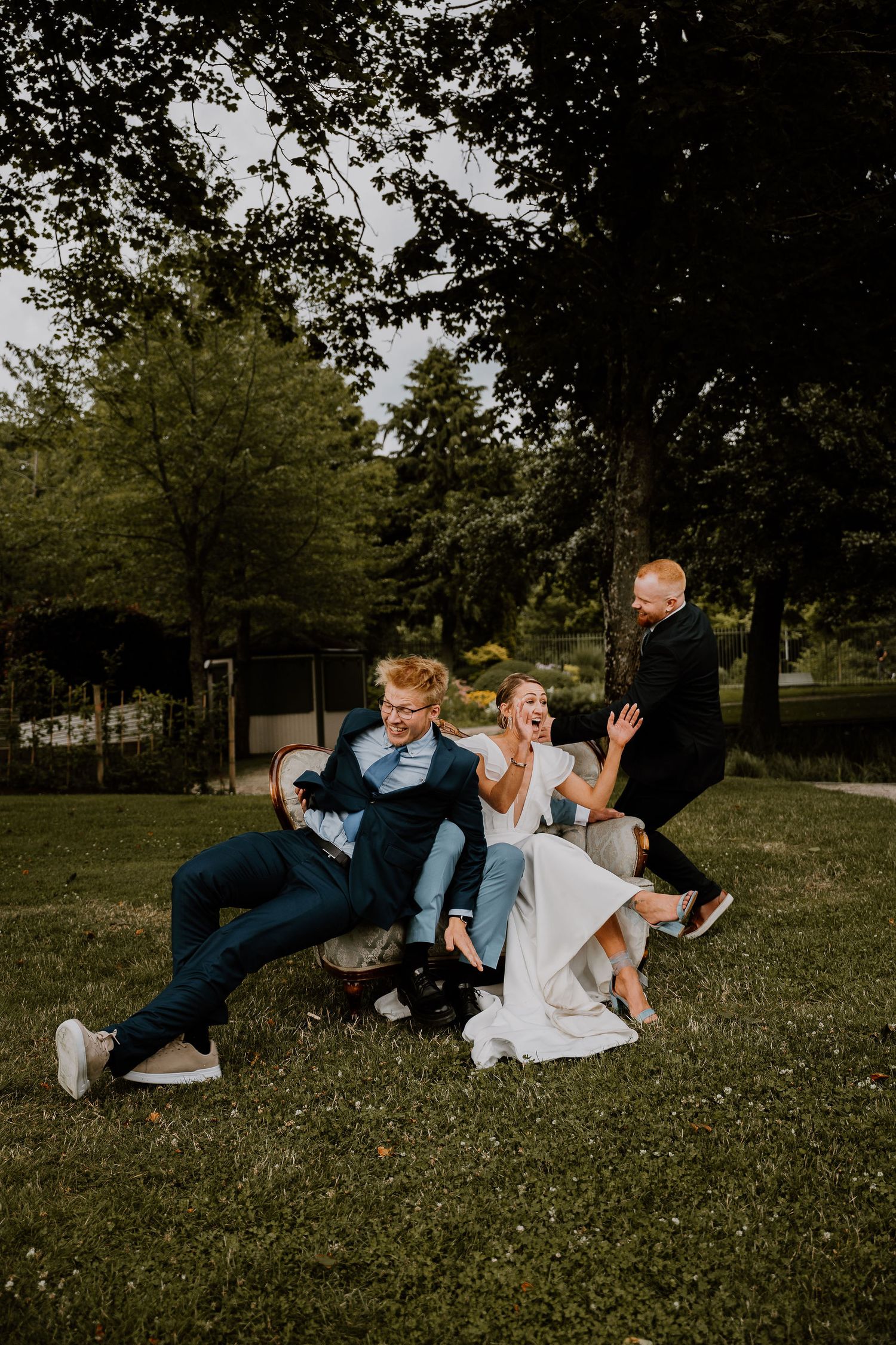 Friends laugh and play while lying on grass during a casual outdoor photoshoot in a park setting.