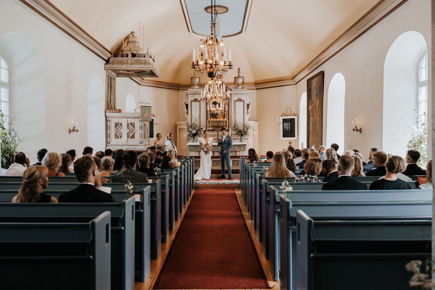 A panoramic view of church wedding guests seated in dark wooden pews under elegant chandeliers during a ceremony.